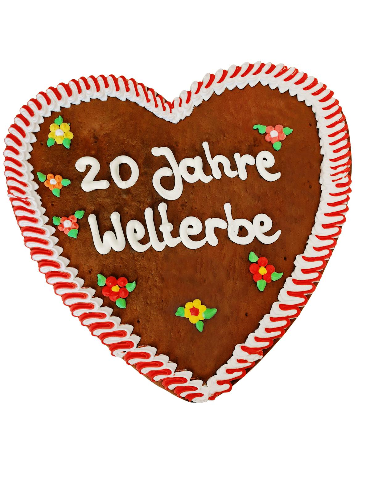 Herzförmiges Lebkuchenherz mit weiß-rotem Zuckerrand und bunten Zuckerdekorationen in Form kleiner Blumen. In der Mitte steht in weißer Zuckerschrift der Schriftzug 20 Jahre Welterbe. Das Herz ist liebevoll verziert und erinnert an traditionelle Jahrmarkt- oder Festtagsgebäcke, gestaltet zur Feier des 20-jährigen Welterbejubiläums.