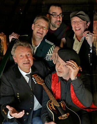 Fünf Männer sitzen und stehen dicht nebeneinander in einem dunklen Raum und lächeln in die Kamera. Einer von ihnen hält eine Gitarre, ein anderer trägt eine Uniformjacke mit Streifen, und ein dritter eine Mütze und Weste mit rotem Glanzstoff. Die Szene wirkt humorvoll und lebendig, als würde es sich um eine Band oder ein Künstlerensemble handeln. Die Beleuchtung hebt ihre Gesichter vor dem dunklen Hintergrund deutlich hervor.