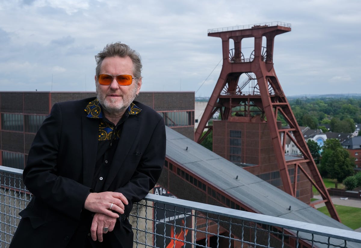 Ein Mann mit grauem Haar, Bart und orangefarbener Sonnenbrille lehnt an einem Geländer auf einer Aussichtsterrasse. Er trägt ein schwarzes Sakko mit einem auffälligen gemusterten Hemdkragen. Im Hintergrund ist das markante Fördergerüst der Zeche Zollverein in Essen zu sehen, flankiert von Backsteingebäuden und grünem Umland unter einem wolkigen Himmel.