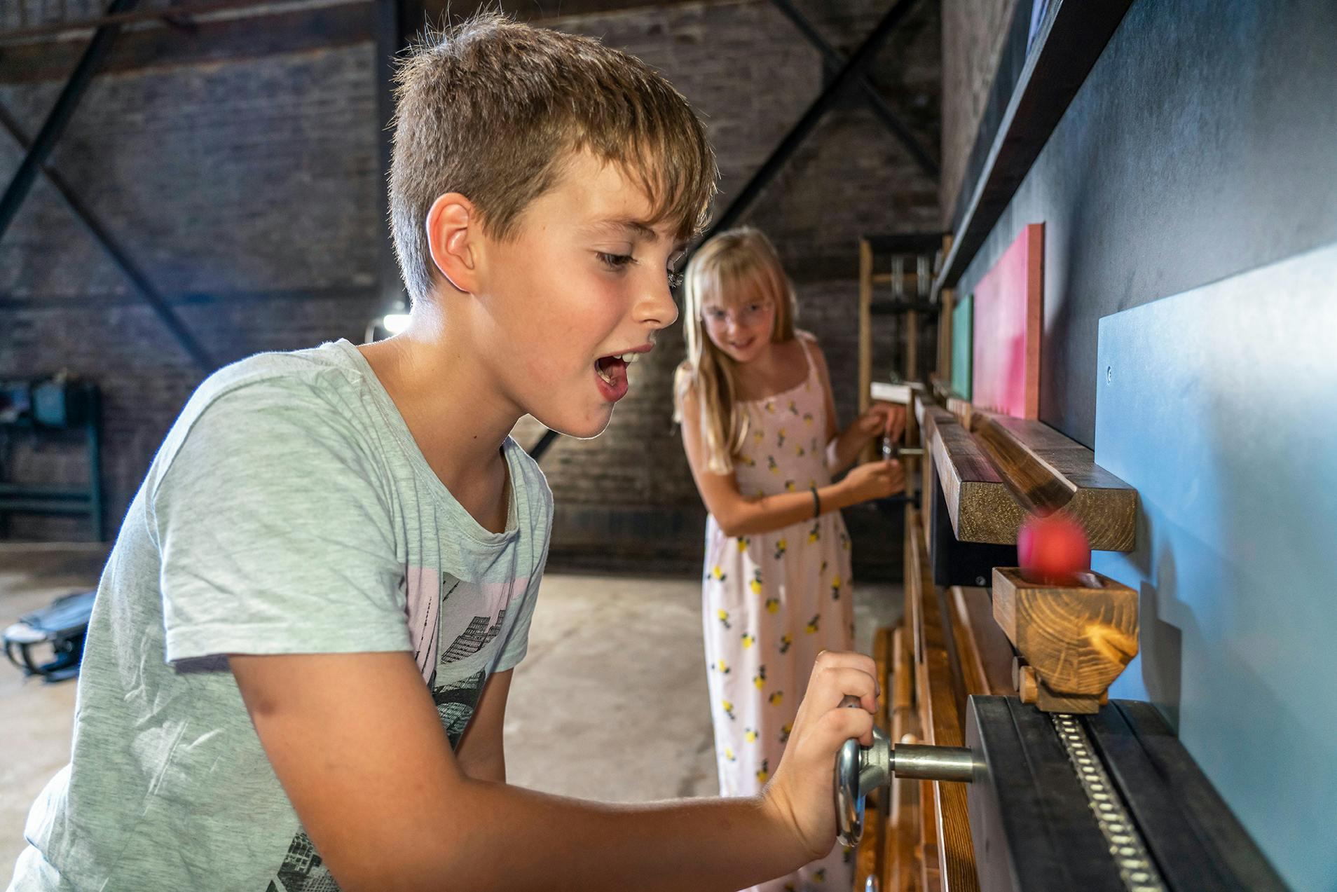Ein Junge im Vordergrund dreht begeistert an einer Kurbel, während eine rote Kugel in einer hölzernen Laufbahn neben ihm rollt. Im Hintergrund ist ein Mädchen zu sehen, das ebenfalls an einem Teil der interaktiven Wandinstallation arbeitet. Beide Kinder befinden sich in einem Raum mit Backsteinwänden und industriellem Charme. Die Szene vermittelt Neugierde und Freude am gemeinsamen Experimentieren.