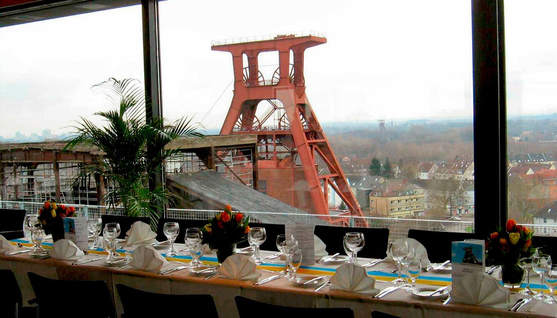 Festlich gedeckter Tisch mit weißen Servietten, Gläsern und Blumenarrangements in einem Raum mit großen Panoramafenstern, durch die man den markanten roten Förderturm der Zeche Zollverein und die umliegende Stadtlandschaft sehen kann. Eine Palme steht als Dekoration im Raum.