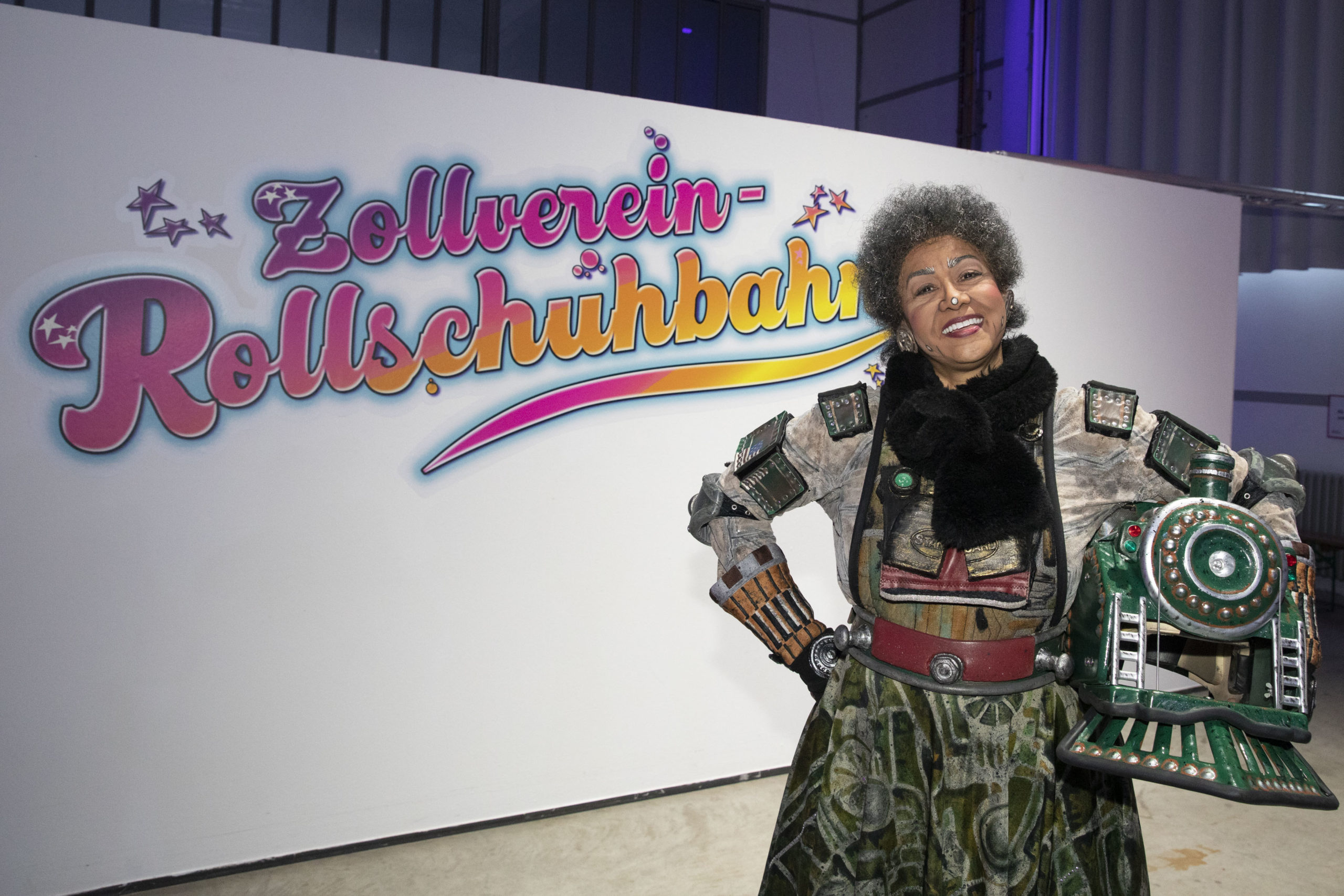 Eine Frau mit lockigem grauem Haar steht lächelnd vor einer Wand mit farbenfrohem Schriftzug Zollverein Rollschuhbahn. Sie trägt ein aufwendig gestaltetes Kostüm im Steampunk-Stil mit Schulterplatten und Armteilen aus Metall sowie einem großen Helm in Form einer Lokomotive an der Hüfte eingeklemmt. Die Pose ist selbstbewusst und freundlich, der Hintergrund ist hell beleuchtet mit einem verspielten Design.