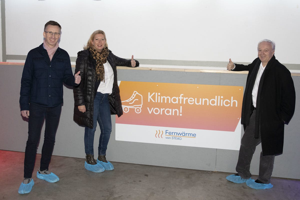 Drei Personen stehen lächelnd nebeneinander vor einem Schild mit der Aufschrift klimafreundlich voran. Alle drei tragen blaue Überzieher über ihren Schuhen. Der Mann links trägt eine dunkle Jeansjacke und eine rosafarbene Schal, die Frau in der Mitte eine schwarze Jacke und einen Leopardenmuster-Schal, der Mann rechts ist in einen dunklen Mantel gekleidet. Sie zeigen Daumen hoch in Richtung Kamera. Das Schild ist orange-gelb verlaufend mit einem Piktogramm eines Rollschuhs und dem Logo Fernwärme von STEAG.