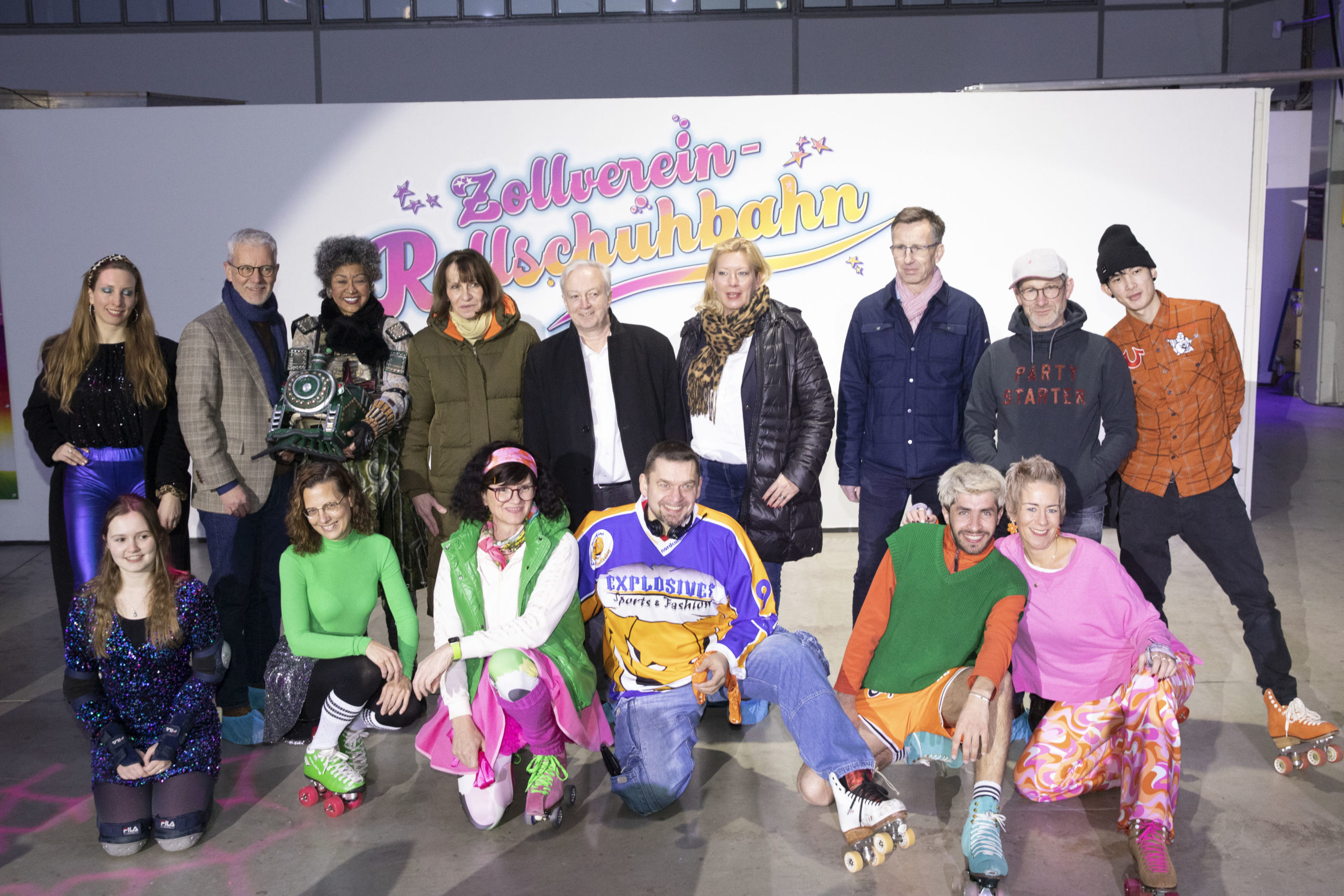 Eine Gruppe von Menschen posiert fröhlich vor einer Wand mit dem Schriftzug Zollverein Rollschuhbahn. Die Personen sind unterschiedlich gekleidet, einige in farbenfrohen Retro-Outfits und Rollschuhen, andere in moderner Alltagskleidung. Eine Frau hält eine futuristisch anmutende Kopfbedeckung in der Hand. Die Stimmung ist lebendig und einladend, mit einem Hauch Nostalgie und Festlichkeit.