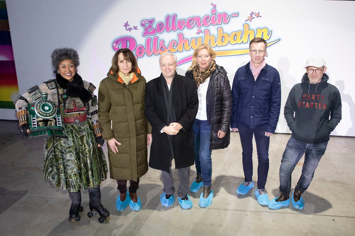Gruppenfoto von sechs Personen, die nebeneinander vor einer weiß beleuchteten Wand mit dem Schriftzug Zollverein-Rollschuhbahn stehen. Die Frau ganz links trägt ein auffälliges Kostüm mit Rollschuhen und einem großen Lokomotiv-Helm im Arm. Die anderen Personen sind winterlich gekleidet und tragen blaue Überzieher über ihren Schuhen. Alle blicken freundlich in die Kamera. Die Szene wirkt wie eine offizielle Eröffnung oder ein Event im Innenbereich.