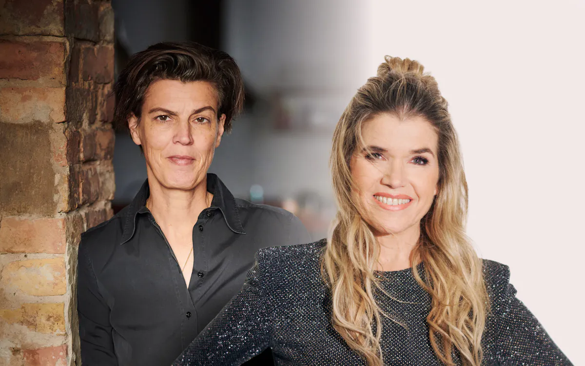 Zwei Frauen stehen nebeneinander in einem Porträtfoto mit geteiltem Hintergrund. Die Frau links hat kurze dunkle Haare, trägt ein schwarzes Hemd und steht vor einer Backsteinwand. Die Frau rechts hat lange, wellige, blonde Haare, trägt ein glitzerndes dunkelgraues Oberteil und lächelt vor einem hellen, unscharfen Hintergrund.