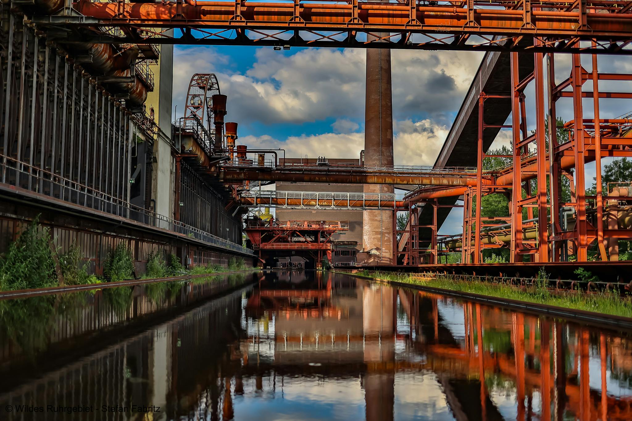 Blick auf ein beeindruckendes Industriedenkmal mit rostroten Stahlkonstruktionen, Rohrleitungen und einer hohen Schornsteinanlage. Die Szene wird durch ein ruhiges Wasserbecken im Vordergrund gespiegelt, was eine symmetrische und atmosphärische Wirkung erzeugt. Im Hintergrund kontrastieren grüne Bäume und ein blauer Himmel mit weißen Wolken die industrielle Architektur. Links und rechts des Wassers verlaufen Gleise und Vegetation.