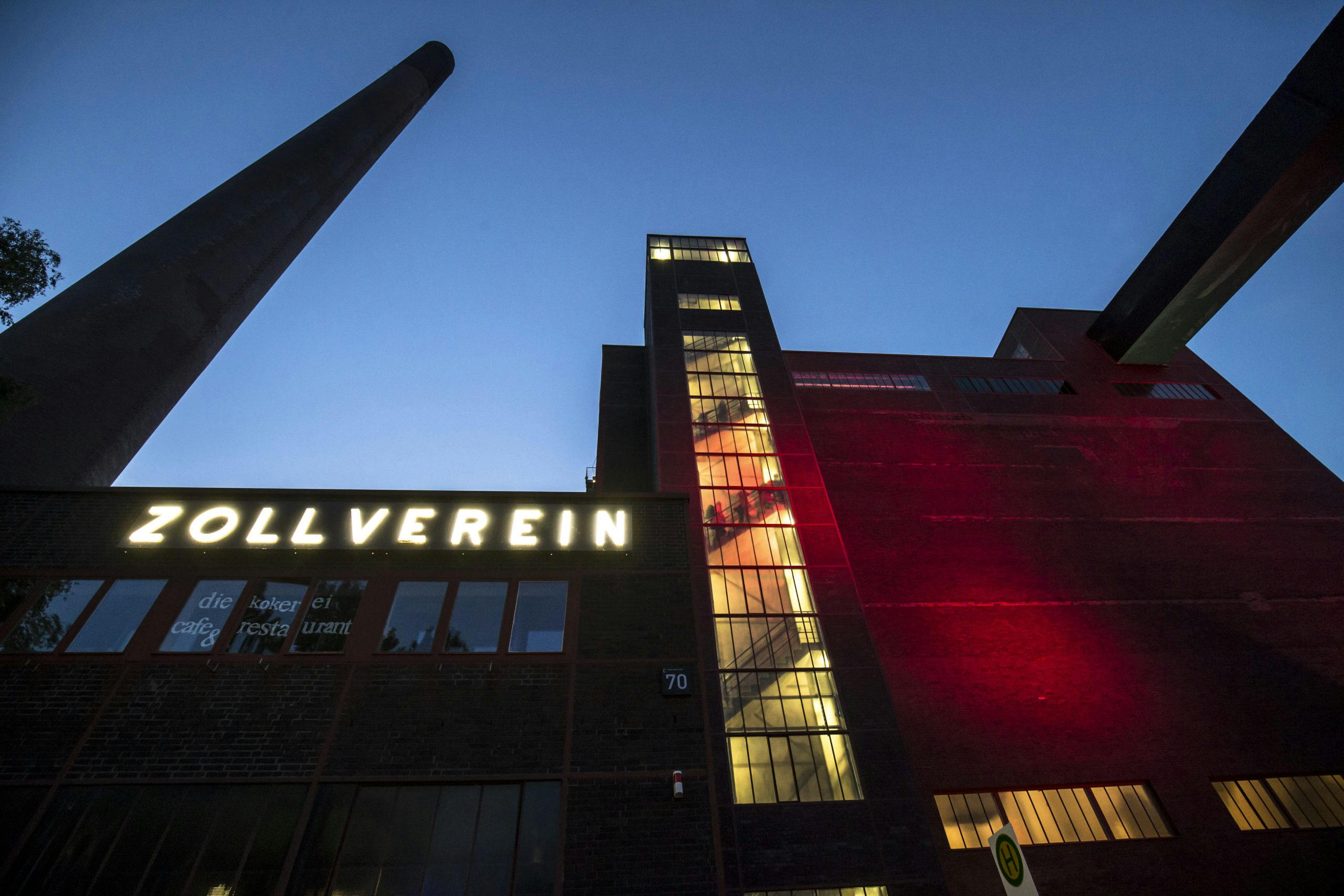 Abendaufnahme des Gebäudes der Kokerei Zollverein mit leuchtender Beschriftung Zollverein an der Fassade. Rechts im Bild verläuft ein markanter Förderbandsteg über das rote Backsteingebäude. Der zentrale Treppenturm ist hell erleuchtet und von innen sichtbar, einige Personen sind auf den Treppen zu erkennen. Links ragt ein hoher Industrieschornstein in den blauen Abendhimmel.