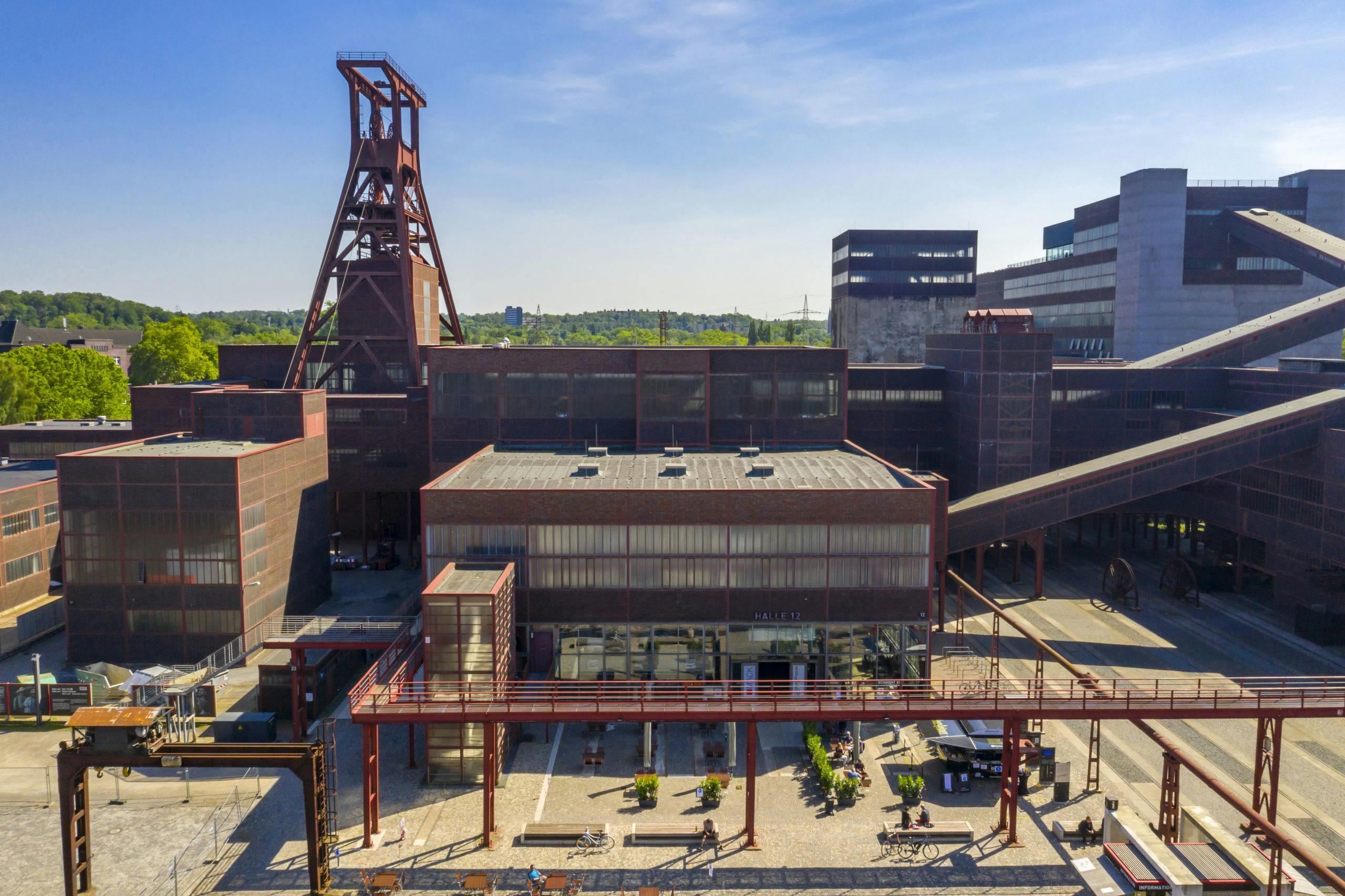 Luftaufnahme des UNESCO-Welterbes Zollverein in Essen an einem sonnigen Tag. Im Mittelpunkt steht die historische Schachtanlage mit dem markanten roten Förderturm über Schacht XII. Die umliegenden Backsteingebäude mit großen Industriefenstern und modernen Anbauten zeugen von der ehemaligen Zeche. Im Vordergrund ist der Vorplatz mit Sitzgelegenheiten, Fahrradständern und Menschen zu sehen, die das Gelände besuchen. Im Hintergrund erstreckt sich grüne Landschaft mit Bäumen und vereinzelten Gebäuden. Der Himmel ist blau und wolkenlos.