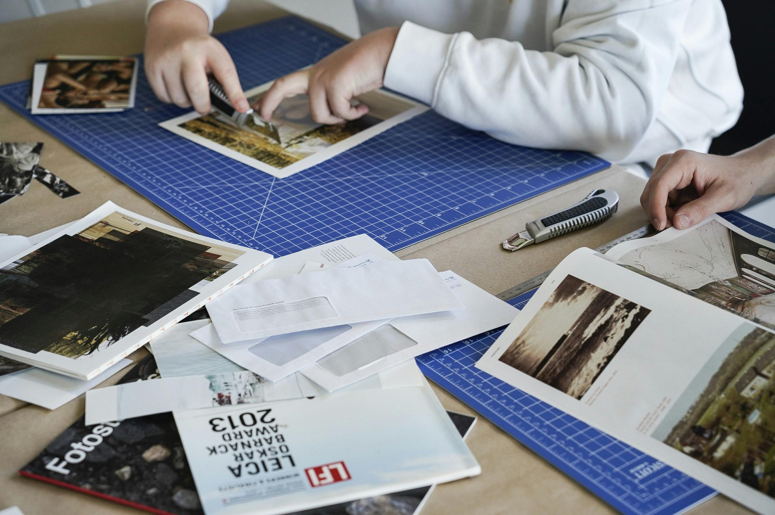 Zwei Personen arbeiten an einem Bastel- oder Designprojekt auf einem Tisch. Sie benutzen Schneidemesser und Lineale auf blauen Schneidematten, um Fotos und Papierbögen zurechtzuschneiden. Um sie herum liegen verschiedene Drucksachen, Zeitschriften, Fotos und Briefumschläge verstreut. Die Szene vermittelt eine kreative Arbeitsatmosphäre.