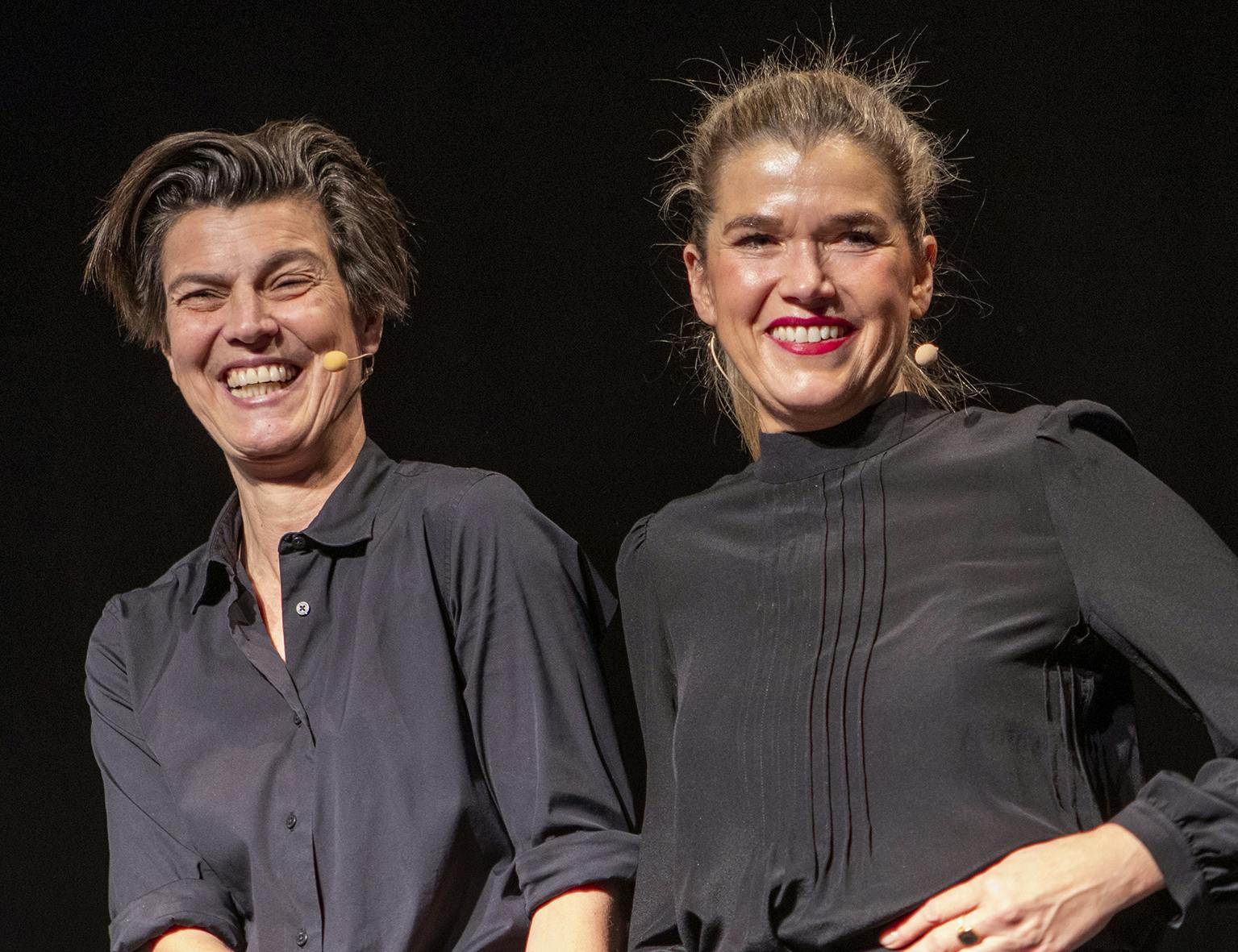 Zwei lachende Frauen stehen nebeneinander auf einer Bühne vor schwarzem Hintergrund. Beide tragen schwarze Oberteile und tragen Headset-Mikrofone. Die Frau links hat kurze, dunkelbraune Haare und ein breites Lächeln, die Frau rechts trägt einen Pferdeschwanz und roten Lippenstift. Sie wirken fröhlich und selbstbewusst.