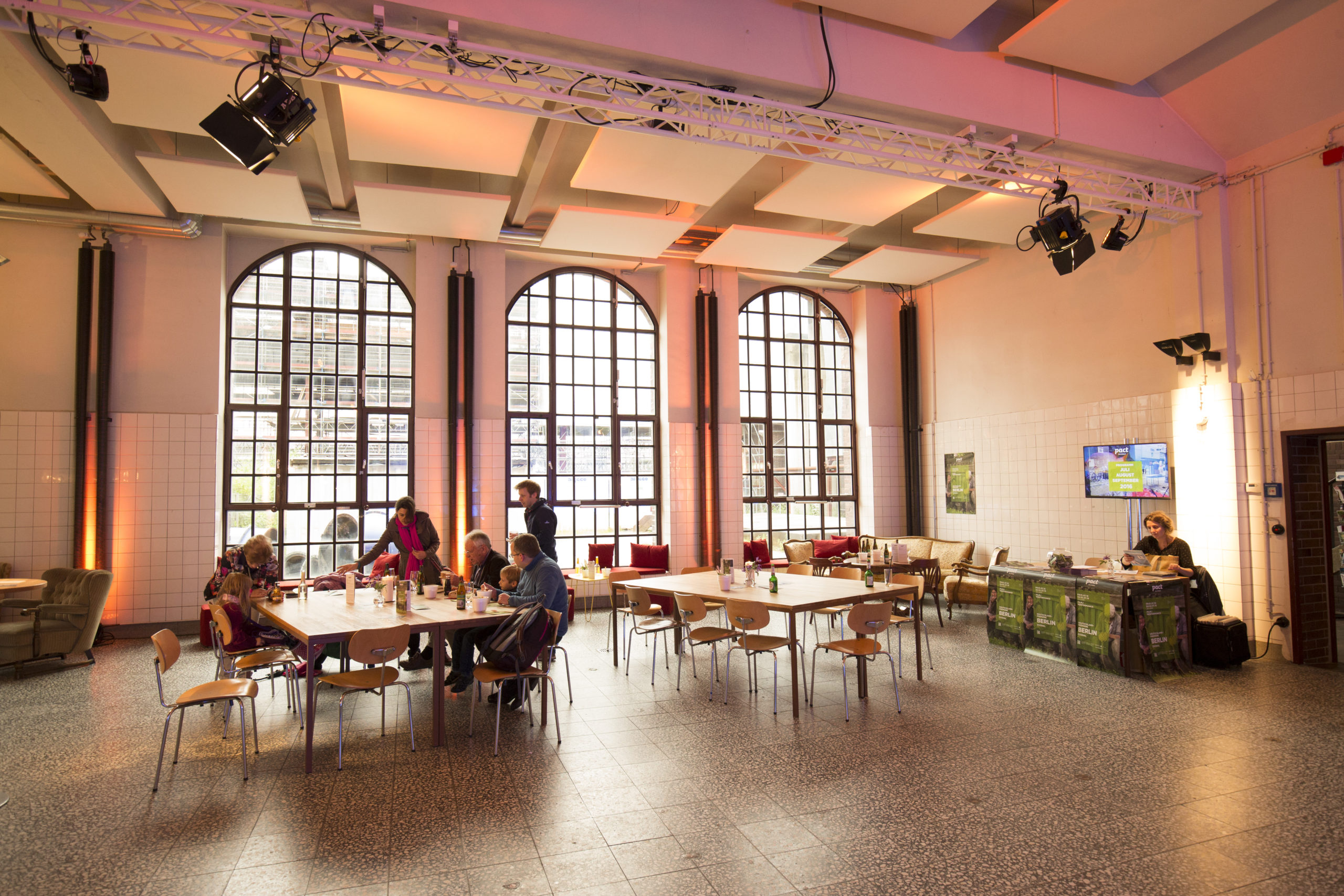 Ein heller, großzügiger Raum mit hoher Decke und großen, bogenförmigen Sprossenfenstern. Mehrere Tische und Stühle stehen locker verteilt im Raum. An einem Tisch in der Mitte sitzen Erwachsene und Kinder, die miteinander essen und sich unterhalten. Links im Raum stehen Sessel, rechts hinten ist eine Sofaecke mit roten Kissen zu sehen. An der rechten Wand befindet sich ein kleiner Tresen mit einer Person dahinter, die etwas schreibt oder liest. Darüber hängt ein Monitor mit Veranstaltungsinformationen. Die Wände sind halbhoch mit weißen Kacheln verkleidet, die Beleuchtung ist warm und stimmungsvoll. Der Raum wirkt wie ein Ort zum Verweilen – ein Café oder ein Begegnungsraum innerhalb einer Veranstaltungslocation.