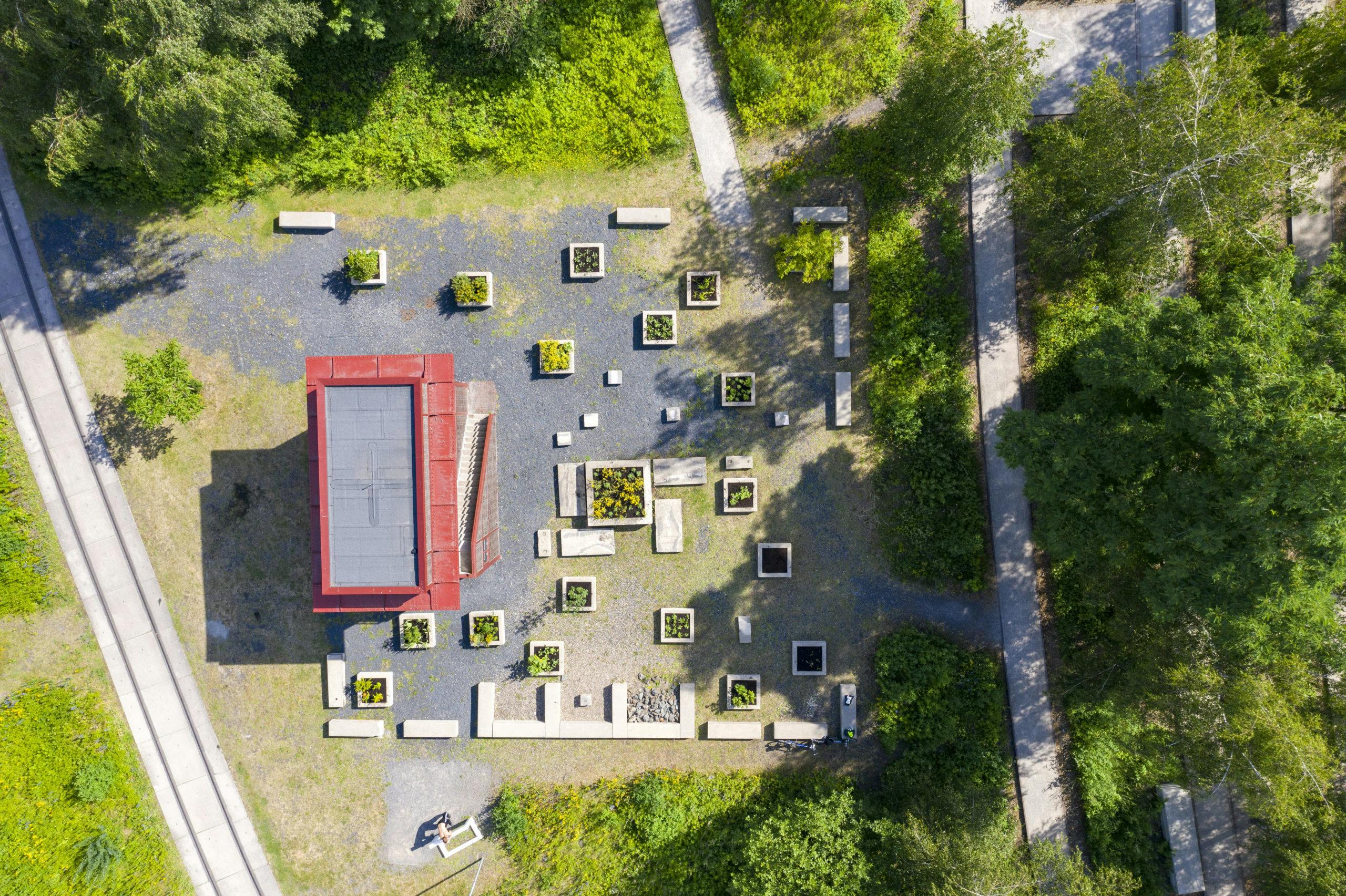 Luftaufnahme eines rechteckigen, roten Pavillons mit Flachdach inmitten eines gestalteten Gartens. Um das Gebäude herum sind zahlreiche quadratische Hochbeete mit unterschiedlichen Pflanzen gleichmäßig angeordnet. Dazwischen stehen helle Betonbänke und -quader, die geometrisch verteilt sind. Wege führen durch das angrenzende grüne Gelände mit Bäumen und Sträuchern. Links ist eine gerade verlaufende Betontrasse zu sehen, für Fußgänger und Radfahrer. Die gesamte Szene wirkt strukturiert und gleichzeitig naturnah.