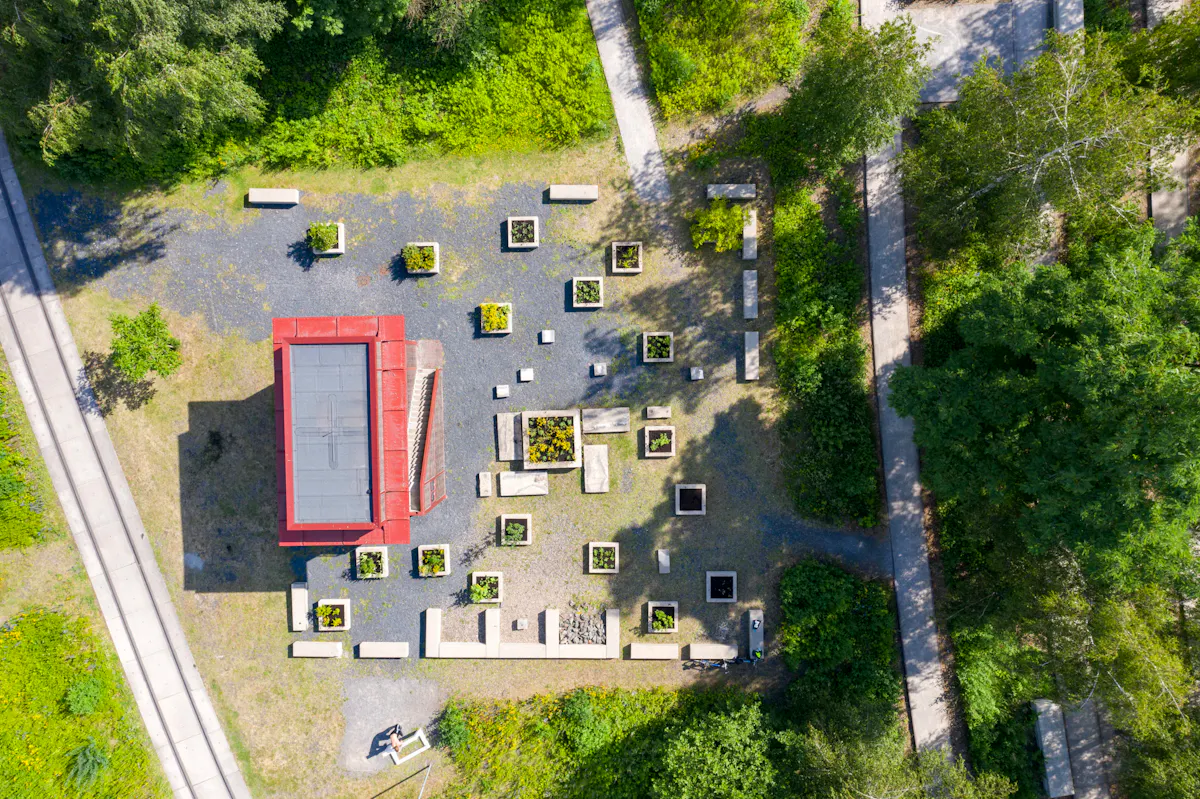 Luftaufnahme eines rechteckigen, roten Pavillons mit Flachdach inmitten eines gestalteten Gartens. Um das Gebäude herum sind zahlreiche quadratische Hochbeete mit unterschiedlichen Pflanzen gleichmäßig angeordnet. Dazwischen stehen helle Betonbänke und -quader, die geometrisch verteilt sind. Wege führen durch das angrenzende grüne Gelände mit Bäumen und Sträuchern. Links ist eine gerade verlaufende Betontrasse zu sehen, für Fußgänger und Radfahrer. Die gesamte Szene wirkt strukturiert und gleichzeitig naturnah.