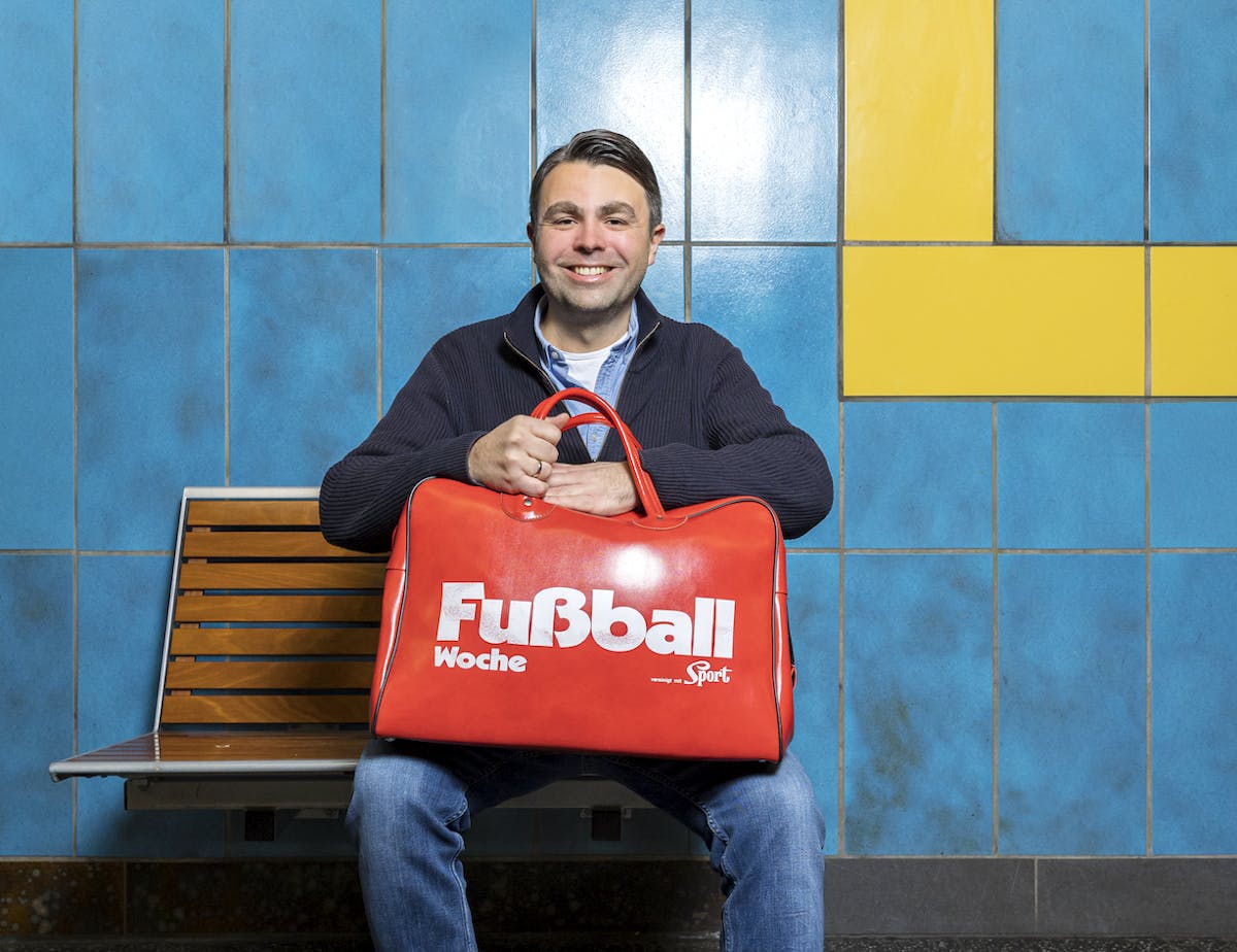 Ein lächelnder Mann sitzt auf einer Holzbank vor einer blau-gelben gefliesten Wand. Er trägt eine blaue Strickjacke und Jeans und hält eine große, leuchtend rote Sporttasche auf dem Schoß. Auf der Tasche steht in weißer Schrift Fußball Woche sowie ein Logo mit dem Wort Sport. Die Szene wirkt freundlich und alltäglich, als ob der Mann auf den nächsten Zug wartet, vielleicht auf dem Weg zum Training oder Spiel.