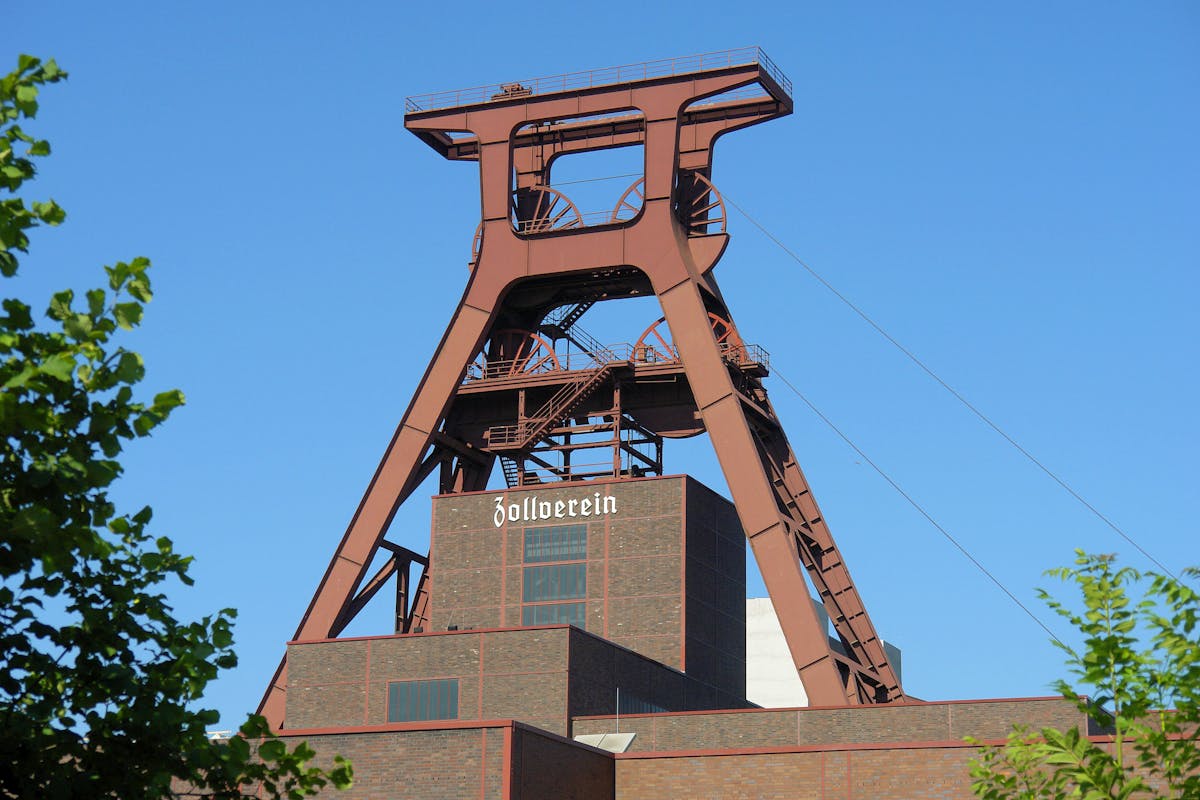 Das Bild zeigt das markante Fördergerüst der Zeche Zollverein in Essen bei strahlend blauem Himmel. Die massive Stahlkonstruktion in rostrot erhebt sich über den Backsteinbau mit der Aufschrift Zollverein. Große Seilscheiben, Treppen und Stahlträger prägen das Bild des ehemaligen Bergwerks, das heute ein Industriedenkmal und UNESCO-Welterbe ist. Im Vordergrund rahmen grüne Blätter von Bäumen das Motiv ein. Die Szene vermittelt industrielle Geschichte in harmonischer Verbindung mit der Natur.