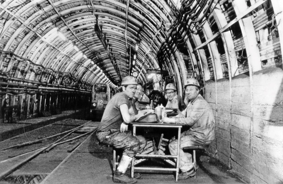 Schwarzweißaufnahme aus einem Bergwerkstunnel. Vier Bergmänner mit Schutzhelmen und schmutziger Arbeitskleidung sitzen an einem einfachen Tisch aus Metall und Holz, offenbar bei einer Pause unter Tage. Im Hintergrund ist ein Grubenfahrzeug mit der Nummer 12 auf den Schienen zu sehen. Der Tunnel ist mit zahlreichen Rohren und Kabeln an den Wänden und der Decke ausgestattet und durch künstliches Licht beleuchtet. Die Szene vermittelt harte körperliche Arbeit und den Alltag im Steinkohlenbergbau.