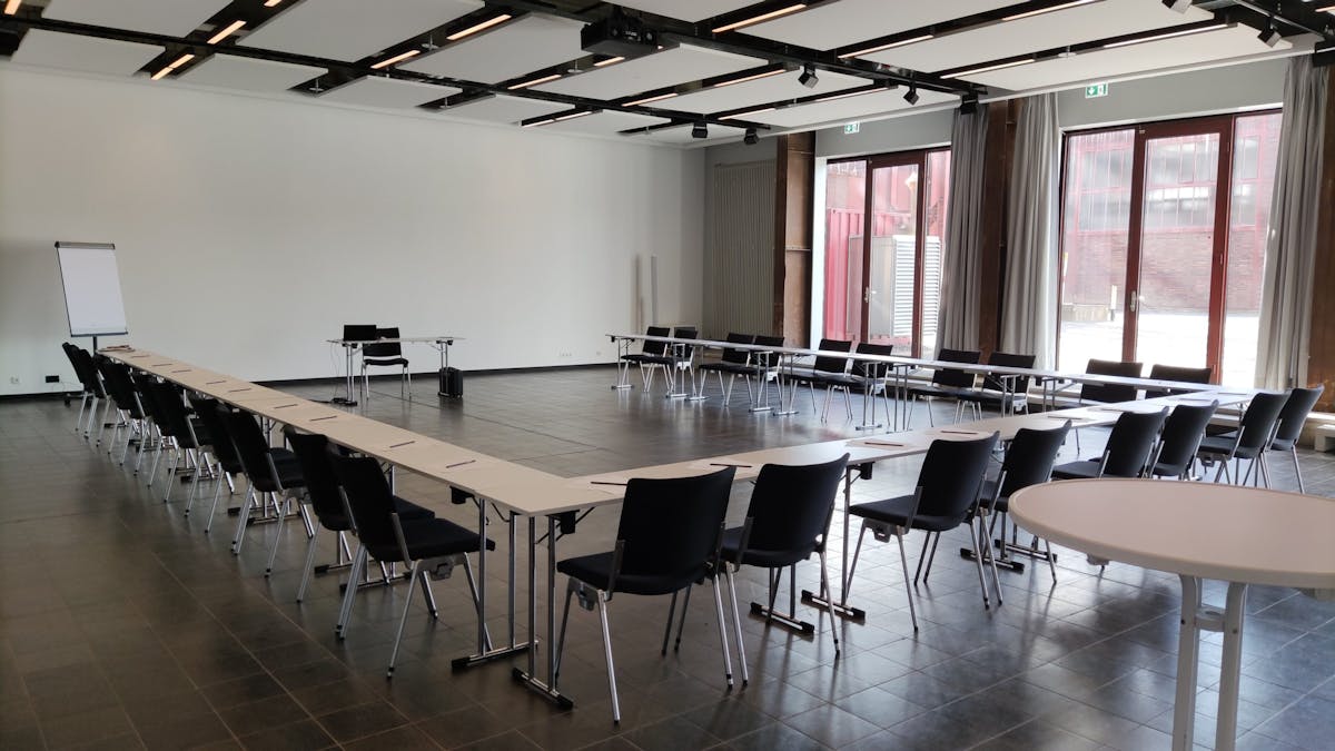 Heller Konferenzraum mit U-förmig angeordneten Tischen und schwarzen Stühlen, vorbereitet für eine Besprechung oder Schulung. Auf den Tischen liegen jeweils Schreibblöcke und Stifte bereit. Vorne steht ein einzelner Tisch mit Laptop und Stuhl sowie ein Flipchart links daneben. Große Fenster mit Vorhängen lassen viel Tageslicht herein und geben den Blick auf das industrielle Außengelände mit Backsteinfassaden frei. Die Atmosphäre wirkt professionell und funktional.