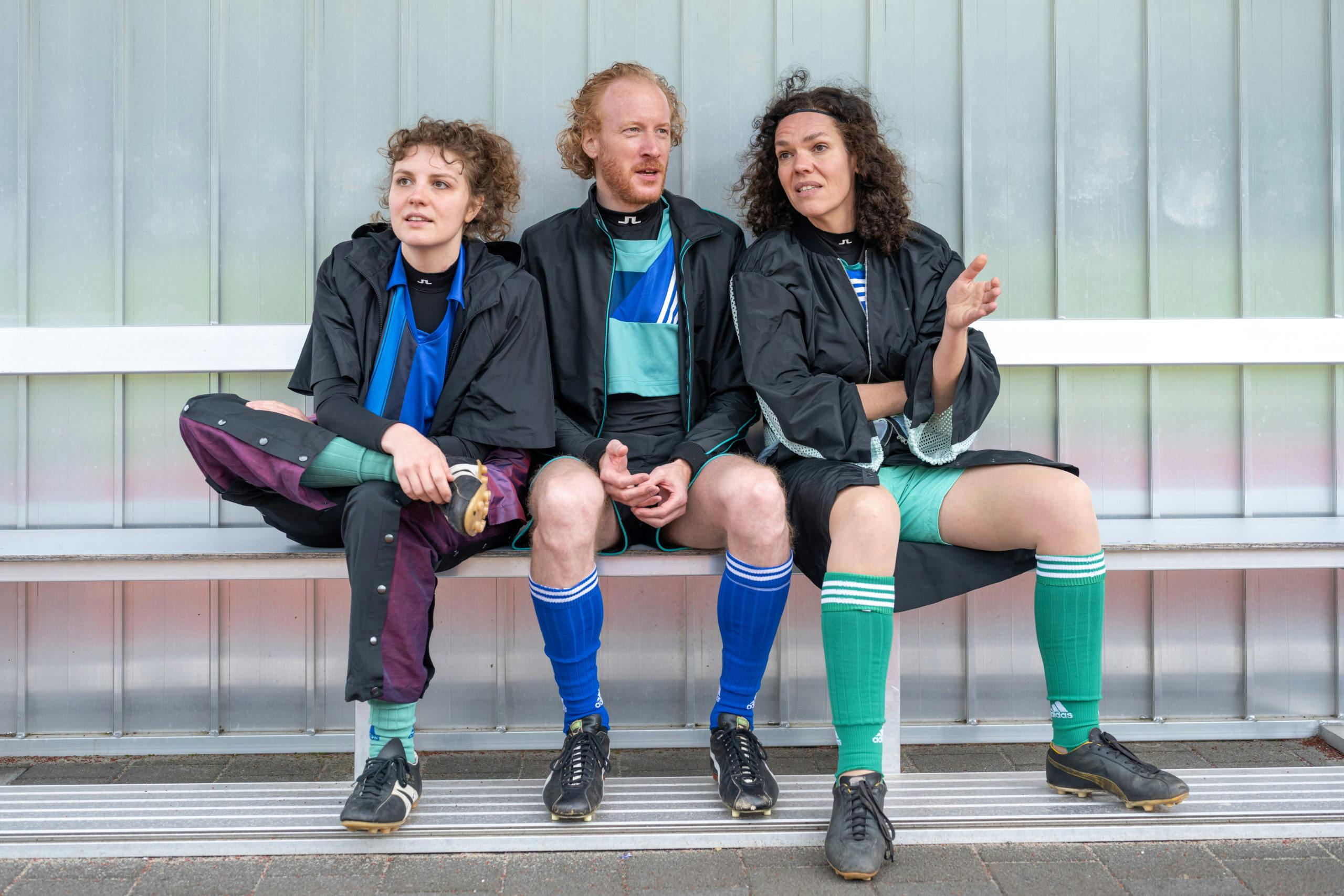 Drei Personen in Sportkleidung sitzen nebeneinander auf einer Ersatzbank vor einer lichtgrauen Wand, vermutlich auf einem Sportplatz. Sie tragen Fußballtrikots, Stutzen und Stollenschuhe in unterschiedlichen Farben sowie schwarze Trainingsjacken. Die Person rechts spricht gestikulierend, während die anderen beiden aufmerksam zuhören. Die Szene wirkt lebendig und authentisch, als befänden sich die Personen in einer Pause oder vor einem Spiel.
