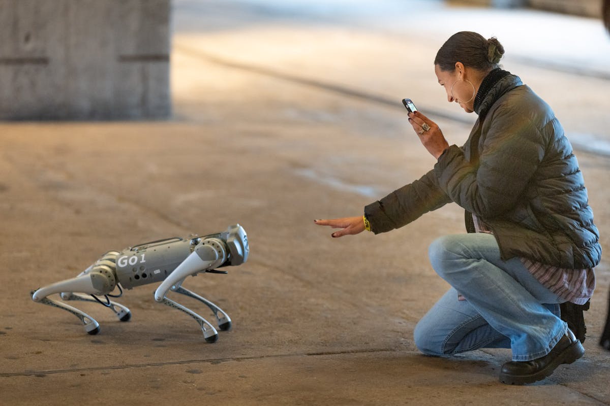 Eine Frau hockt auf dem Boden und interagiert lächelnd mit einem kleinen vierbeinigen Roboter mit der Aufschrift G01. Sie hält ein Smartphone in der Hand, mit dem sie den Roboter fotografiert oder filmt, während sie mit der anderen Hand eine Geste in seine Richtung macht. Die Szene spielt sich in einer offenen, leicht industriell wirkenden Halle oder einem überdachten Außenbereich ab.