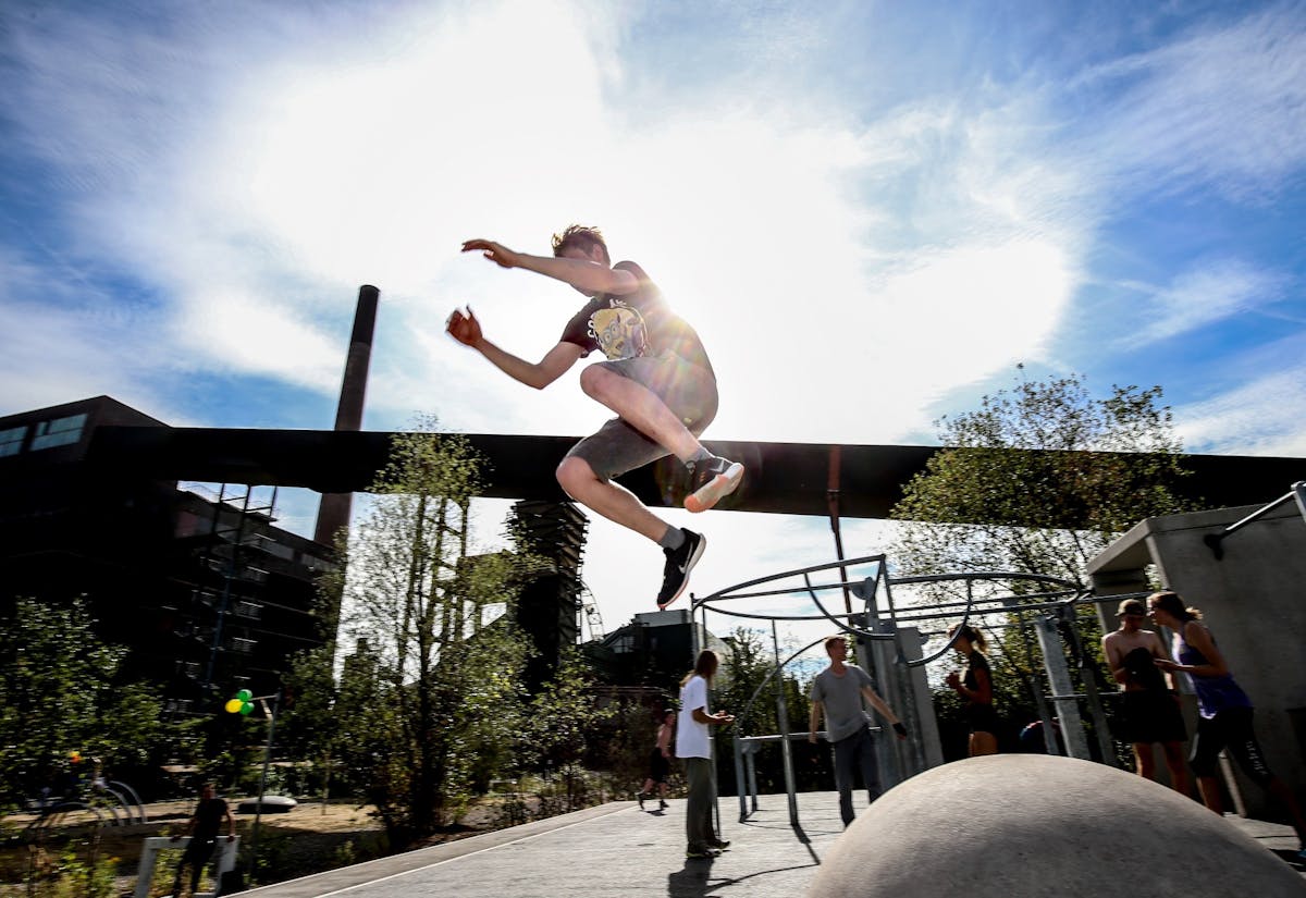 Ein junger Mann springt hoch in die Luft und ist dabei, einen Parkour-Trick auszuführen. Er trägt ein schwarzes T-Shirt mit einem Minions-Motiv, schwarze Shorts und schwarze Sportschuhe mit weißen Sohlen. Die Sonne scheint hell von hinten, wodurch er als Silhouette vor einem strahlend blauen Himmel mit einigen Wolken erscheint. Im Hintergrund sind Bäume, Industriegebäude mit Schornsteinen und eine Gruppe von Menschen zu sehen, die sich unterhalten und die Szene beobachten. Das Bild vermittelt Dynamik, Energie und ein urbanes Lebensgefühl.