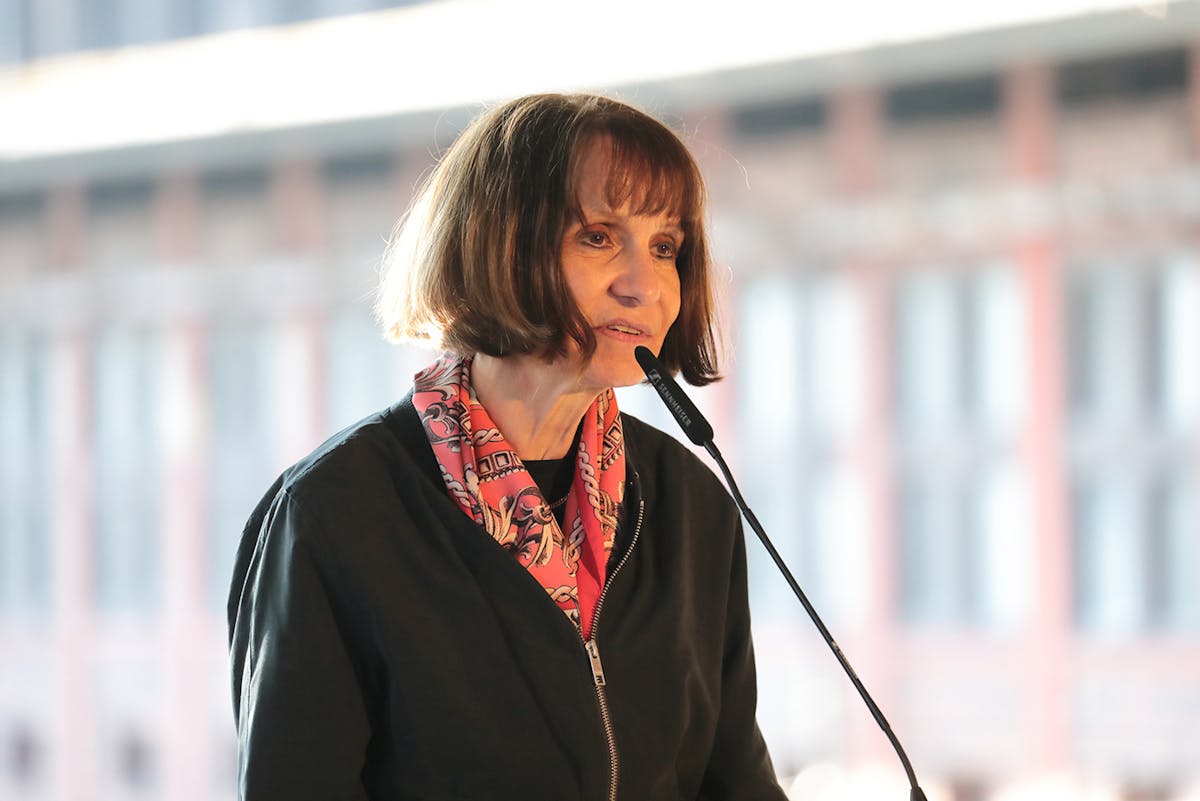 Eine Frau mit kinnlangem braunem Haar und Pony spricht in ein Mikrofon. Sie trägt eine schwarze Jacke und ein gemustertes Halstuch in Rottönen. Der Hintergrund ist unscharf und zeigt eine lichtdurchflutete Industriehalle. Die Szene wirkt wie ein öffentlicher Vortrag oder eine offizielle Rede.