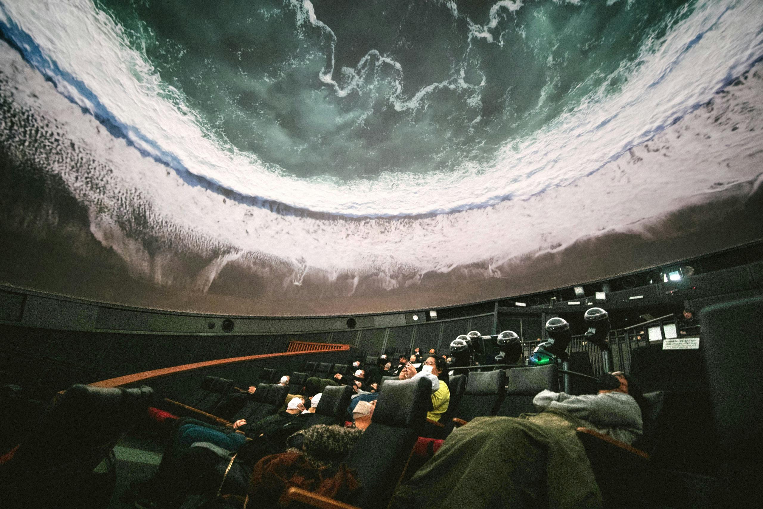Besucherinnen und Besucher sitzen in einem Kuppelraum auf Liegestühlen und betrachten eine beeindruckende Projektion einer Meeresbrandung an der Decke, die realistisch über ihren Köpfen verläuft und ein immersives Naturerlebnis vermittelt.