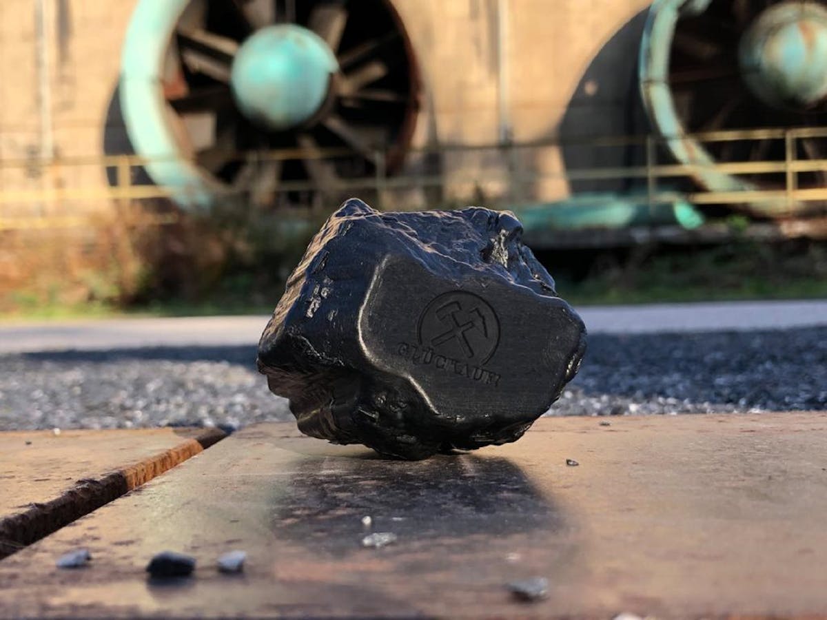 Ein schwarzes Stück Seife in Form eines Kohlestücks liegt auf einer metallischen Oberfläche. Auf der Seife ist ein rundes Logo mit zwei gekreuzten Bergbauschlägeln und dem Wort Glückauf darunter eingeprägt. Im Hintergrund unscharf sind große, runde Lüfter zu erkennen, die Teil eines Industriegebäudes sind. Das Licht der Sonne fällt seitlich auf die Szene und erzeugt Schatten und Glanzlichter auf der Kohle und der Oberfläche.
