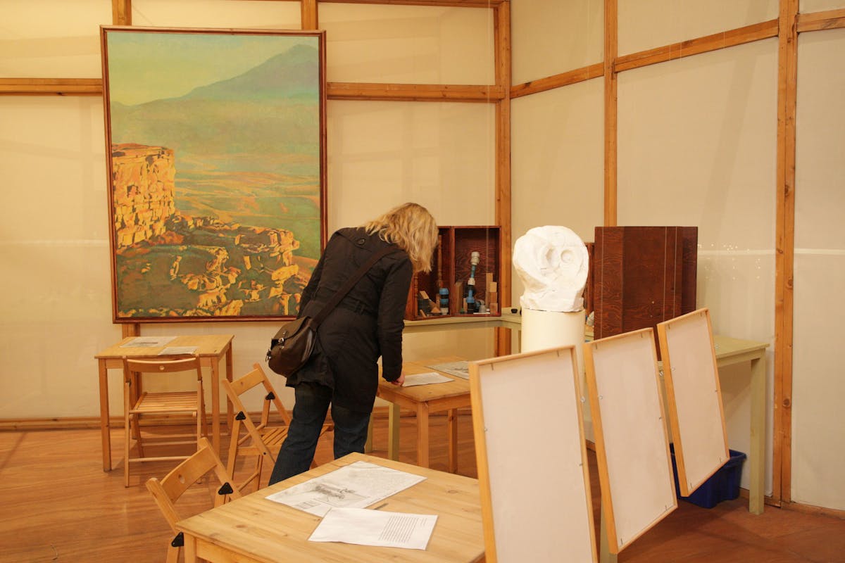 Das Bild zeigt einen Ausstellungsraum mit einer Frau, die an einem Tisch steht und ein Blatt Papier betrachtet. Im Hintergrund hängt ein Gemälde eines Gebirgsmassivs mit hellen, goldenen Farben. Auf den Tischen stehen verschiedene Ausstellungsobjekte, darunter ein Skulpturenabdruck eines Kopfes, und an den Wänden sind Holzrahmen mit leeren Ausstellungsflächen zu sehen. Die Wände des Raumes bestehen aus transparenten, hellen Paneelen und der Boden ist aus Holz. Es herrscht eine ruhige und sachliche Atmosphäre.