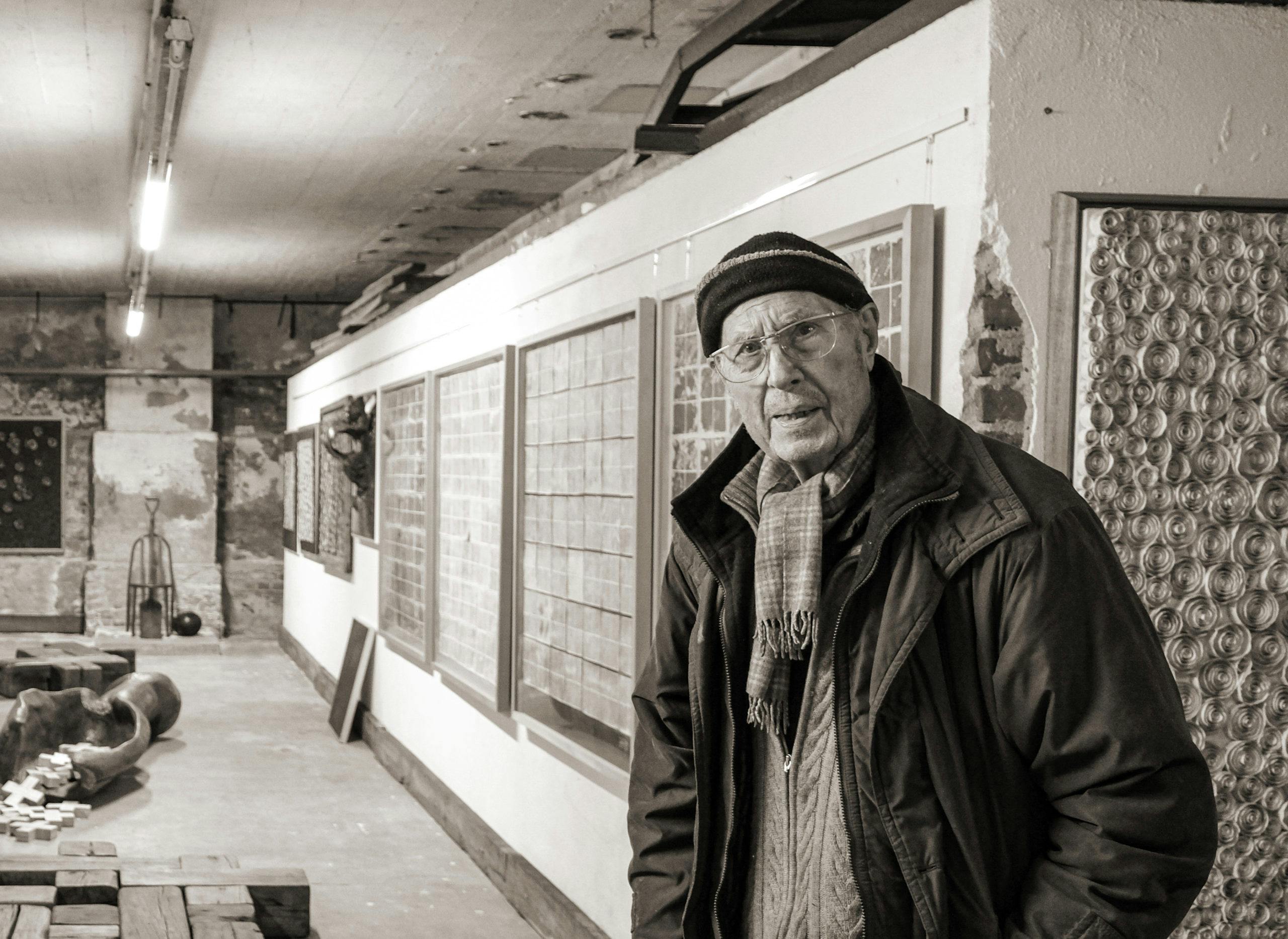 Das Bild zeigt einen älteren Mann in einem dunklen Mantel, einer Mütze und einem Schal, der leicht zur Seite schaut. Er steht in einem Raum mit mehreren Kunstwerken an den Wänden. Im Hintergrund sind mehrere Bilder und Skulpturen zu sehen, darunter ein großes, rundes Kunstwerk und eine Wand mit quadratischen Mustern. Der Raum hat eine industrielle Atmosphäre, mit sichtbaren Ziegelwänden und einem leichten Lichtschein von der Decke.