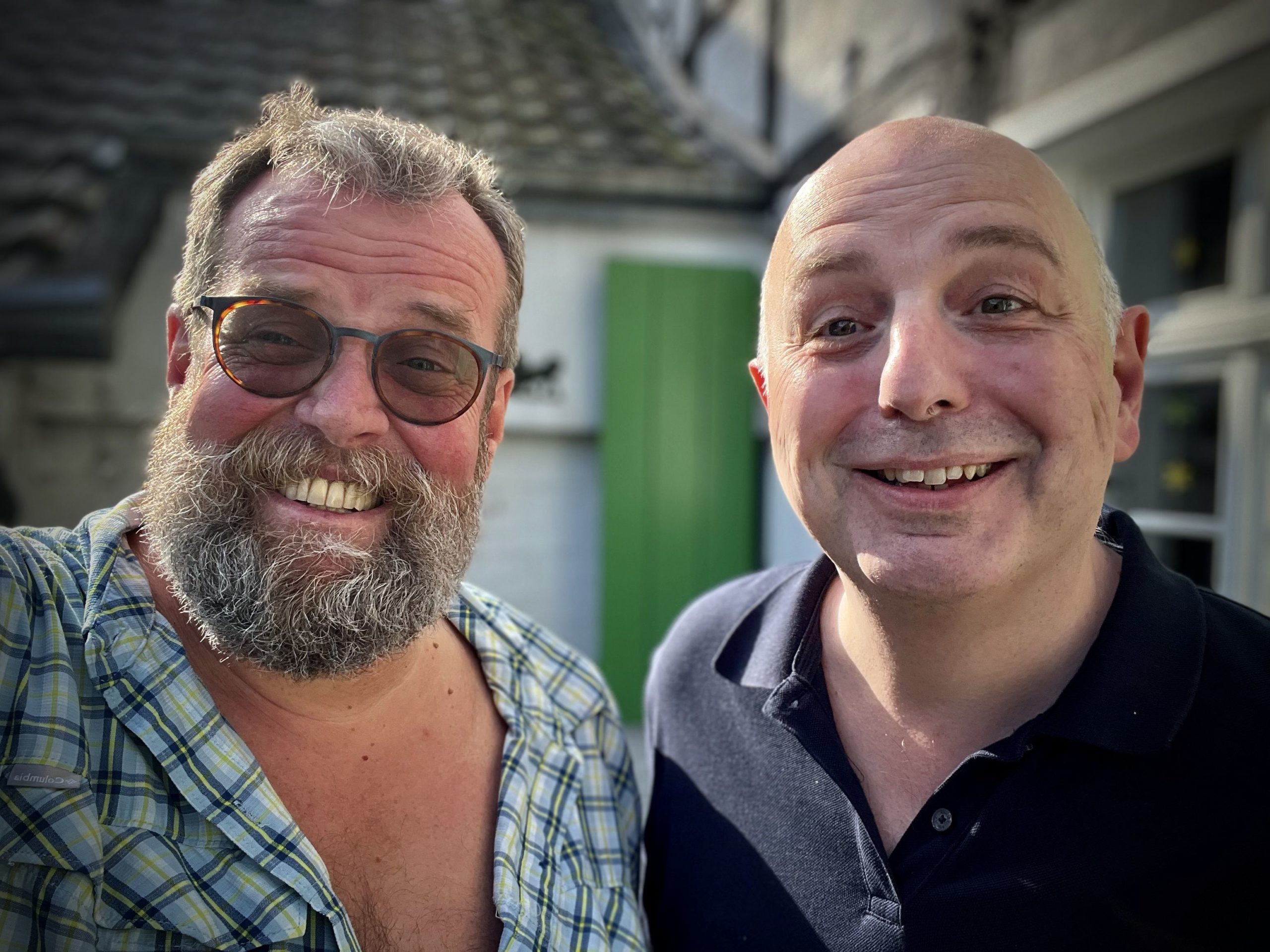 Das Bild zeigt zwei Männer, die zusammen in die Kamera lächeln. Der Mann auf der linken Seite hat einen dichten grauen Bart und trägt eine kariert gemusterte Hemdjacke, die offen getragen wird. Der Mann auf der rechten Seite ist kahl und trägt ein dunkelblaues Polo-Shirt. Beide haben Brillen und strahlen freundlich. Der Hintergrund zeigt Teile eines Hauses mit grünen Fensterläden.