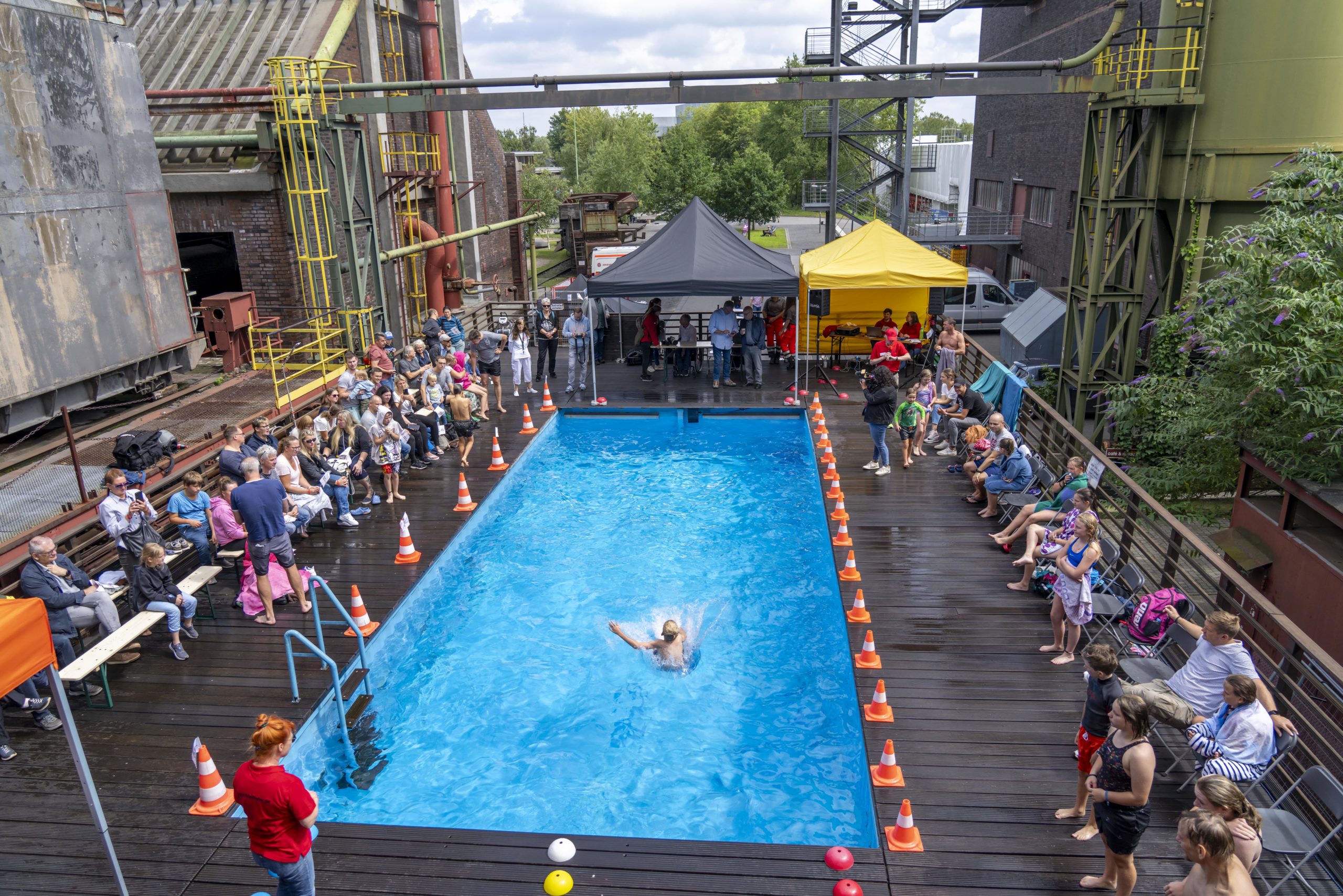 Viele Menschen sitzen und stehen auf einer Holzplattform um ein kleines, rechteckiges blaues Schwimmbecken. Ein Mensch schwimmt gerade in der Mitte des Beckens, eine Person taucht auf der linken Seite aus dem Wasser. Rund um das Becken sind orange-weiße Verkehrskegel aufgestellt. Im Hintergrund stehen zwei Zelte, ein schwarzes und ein gelbes, unter denen sich einige Menschen aufhalten. Die Szenerie ist umgeben von alten Industrieanlagen und grüner Vegetation. Der Himmel ist teilweise bewölkt.
