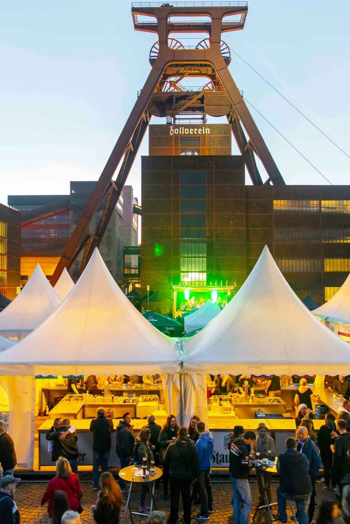 Das Bild zeigt ein belebtes Abendfest auf dem Gelände des UNESCO-Welterbes Zeche Zollverein in Essen. Im Hintergrund ragt das markante Fördergerüst mit der Aufschrift Zollverein über die historischen Backsteingebäude empor. Im Vordergrund stehen mehrere weiße Zelte mit Verkaufsständen, an denen Speisen und Getränke angeboten werden. Davor versammeln sich viele Menschen in Gruppen, einige stehen an Stehtischen mit Getränken in der Hand. Die Szenerie ist durch farbige Lichter stimmungsvoll beleuchtet, im Hintergrund auf einer kleinen Bühne ist grün beleuchtete Livemusik zu sehen. Die Atmosphäre wirkt lebendig und gesellig.