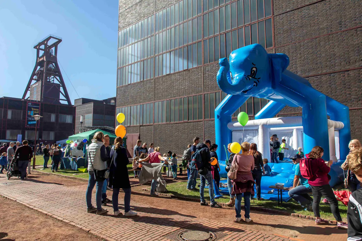 Viele Menschen, darunter Kinder und Erwachsene, versammeln sich an einem sonnigen Tag draußen auf einem Gelände mit Industriebauten im Hintergrund. Rechts im Bild steht eine große blaue Hüpfburg in Form eines Elefanten, um die sich Kinder und Erwachsene gruppieren. Einige Menschen halten bunte Luftballons. Im Hintergrund ist eine Industrieanlage mit einem Fördergerüst zu sehen. Die Stimmung wirkt lebhaft und fröhlich.