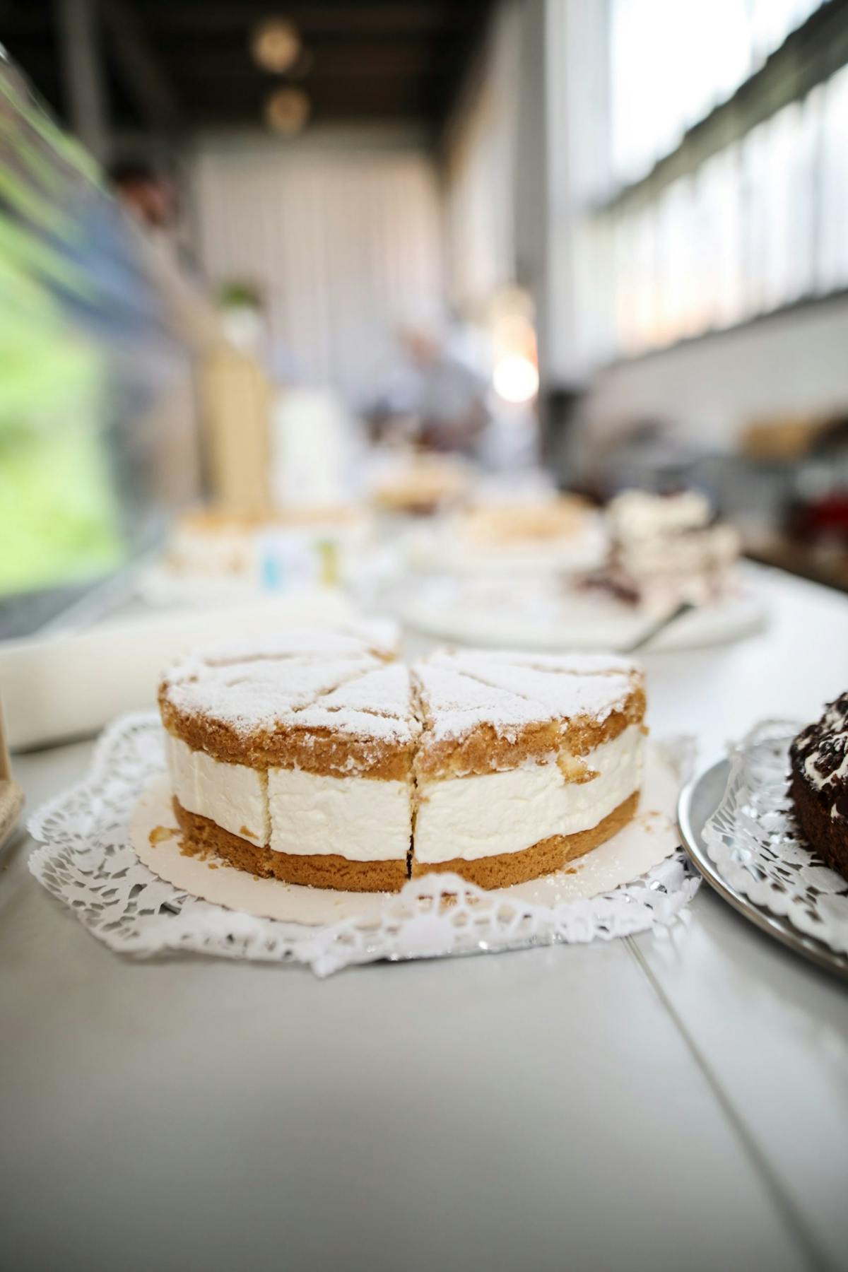 Auf dem Bild ist eine Sahnetorte mit mehreren Stücken auf einer weißen Tortenspitze zu sehen. Die Torte besteht aus einem hellbraunen Boden, einer dicken Schicht Sahne und einem weiteren Teigboden, der mit Puderzucker bestreut ist. Sie steht auf einem gedeckten Tisch in einer hellen Umgebung mit großen Fenstern, durch die Tageslicht einfällt. Im Hintergrund sind weitere Kuchen und unscharfe Personen zu erkennen, was auf eine Veranstaltung oder ein Café hindeutet. Rechts im Bildrand ist ein weiterer dunklerer Kuchen angeschnitten zu sehen. Die Atmosphäre wirkt freundlich und einladend.