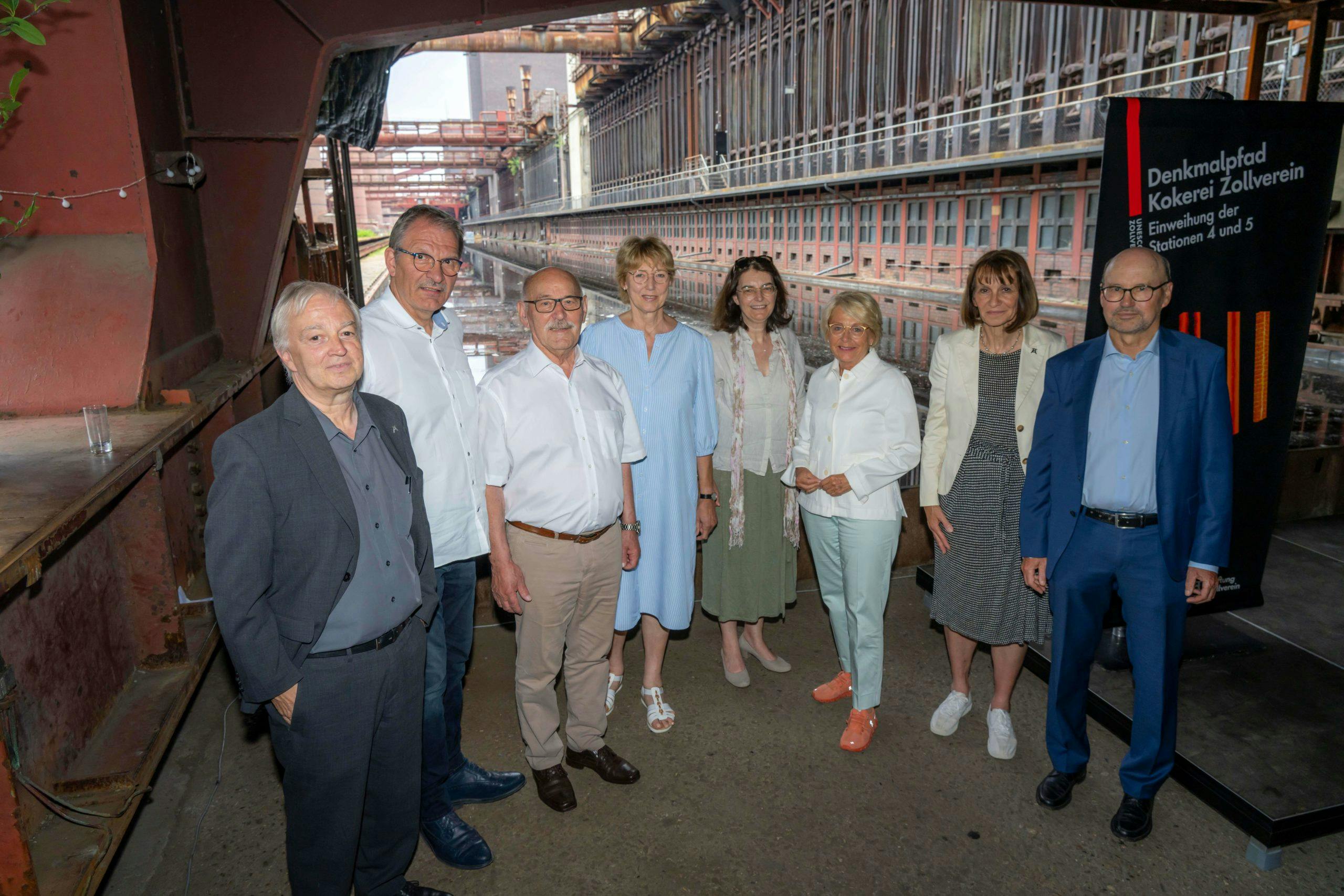 Eine Gruppe von neun Personen steht unter einer Stahlbrücke oder einem großen Stahlträger in einem Industriegebiet. Die Personen sind locker nebeneinander aufgereiht und blicken in die Kamera. Sie tragen überwiegend elegante Kleidung, mit Hemden, Blusen, Anzügen und Kleidern in gedeckten Farben. Im Hintergrund ist ein langer, schmales Wasserbecken zu sehen, das von einer alten Industrieanlage mit vielen Fenstern und Stahlrohren umgeben ist. Rechts neben der Gruppe steht ein schwarzes Schild mit der Aufschrift Denkmalpfad Kokerei Zollverein Einweihung der Stationen 4 und 5. Die Stimmung wirkt offiziell und freundlich, vermutlich im Rahmen einer Veranstaltung oder Einweihung.