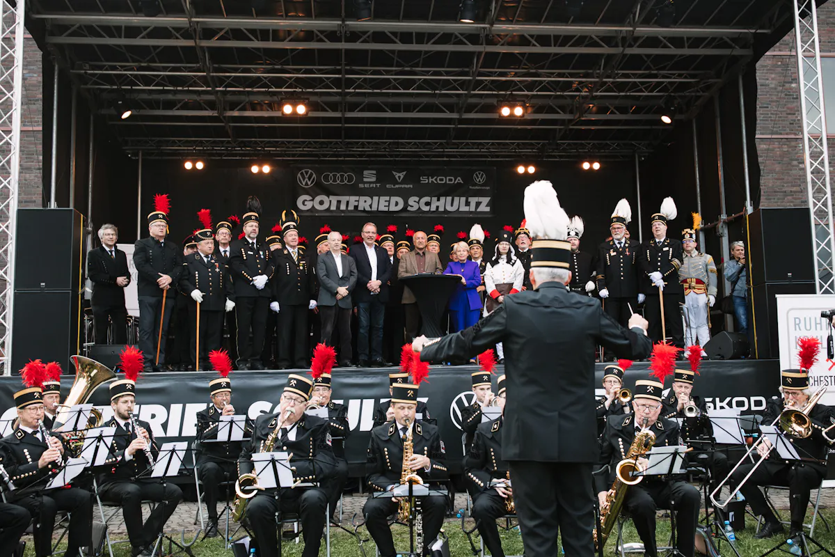 Das Bild zeigt eine festlich geschmückte Bühne mit einer großen Gruppe von Menschen in historischer Bergmannsuniform. Die Uniformierten tragen schwarze Jacken mit goldenen Knöpfen und Helme mit weißen oder roten Federbüschen. In der Mitte der Bühne stehen auch einige Personen in ziviler Kleidung, darunter eine Frau in einem auffälligen blauen Mantel. Vor der Bühne spielt eine Bergmannskapelle mit Blasinstrumenten wie Trompeten, Saxophonen und Tuben, dirigiert von einem Mann mit weißem Federbusch auf dem Helm. Über der Bühne hängen Sponsorenlogos, unter anderem von Volkswagen, Audi, Škoda und dem Autohaus Gottfried Schultz. Die Veranstaltung wirkt wie ein traditionelles Bergfest mit Musik und Ehrenformation.