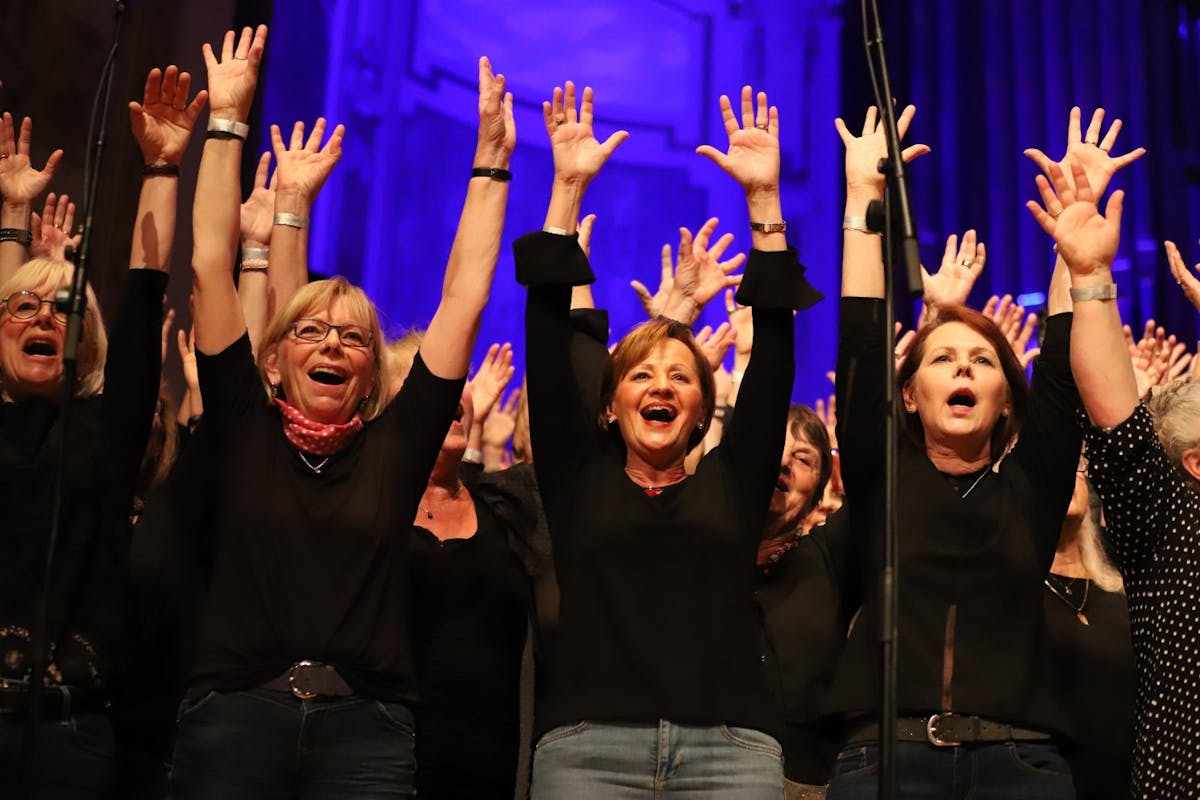 Mehrere Frauen eines Chors stehen eng beieinander vor einem Hintergrund mit violett-blauem Bühnenlicht. Sie tragen überwiegend schwarze Kleidung und haben die Arme nach oben gestreckt. Sie singen mit ausdrucksstarken Mienen und wirken sehr engagiert und fröhlich. Einige tragen Brillen und Schmuck, manche haben kurze, andere lange Haare. Ein Mikrofonständer ist im Vordergrund sichtbar.