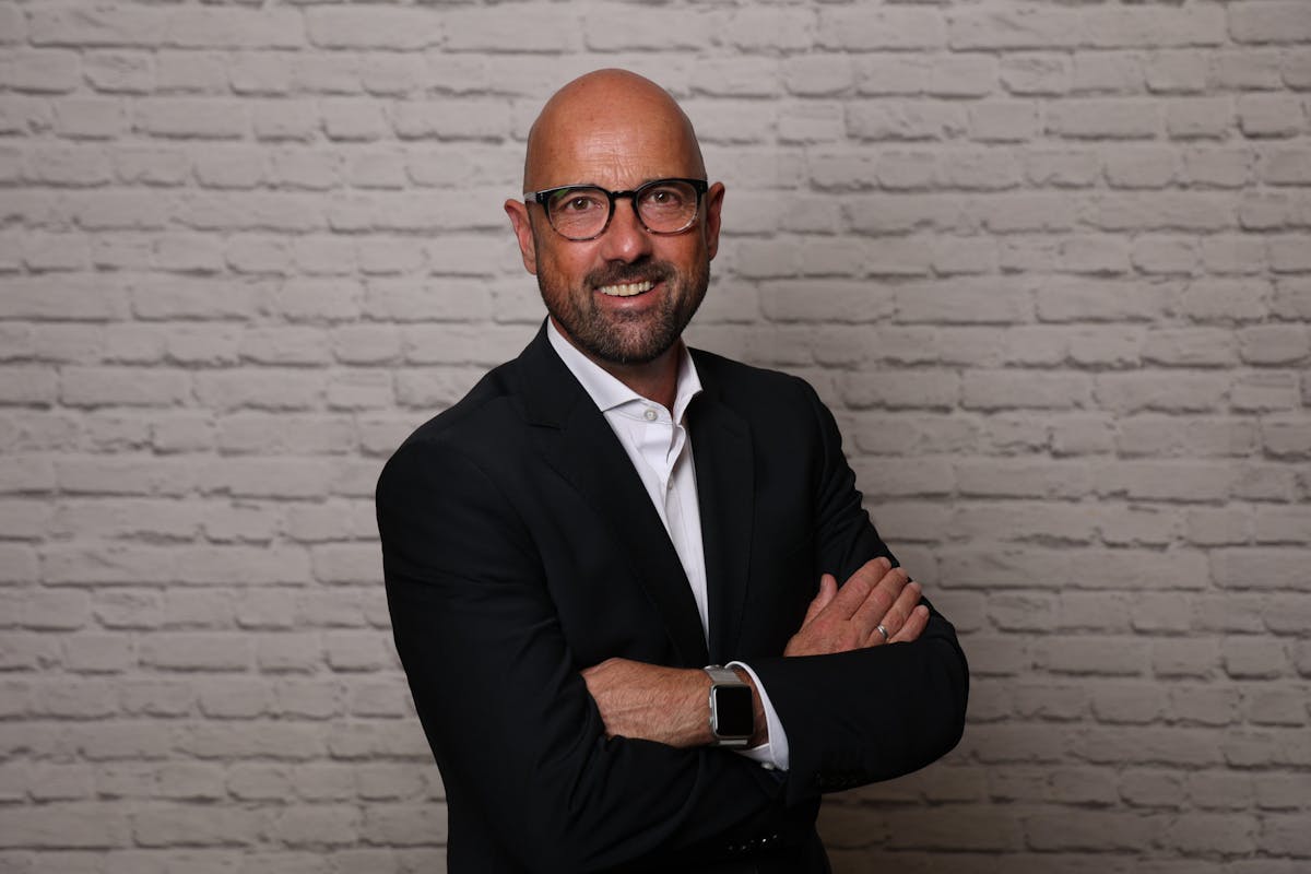 Mann mit Glatze und Vollbart steht vor einer weißen Ziegelsteinwand, trägt eine schwarze Brille, ein schwarzes Sakko über einem weißen Hemd, und lächelt mit verschränkten Armen in die Kamera. An seinem linken Handgelenk ist eine Smartwatch zu sehen.