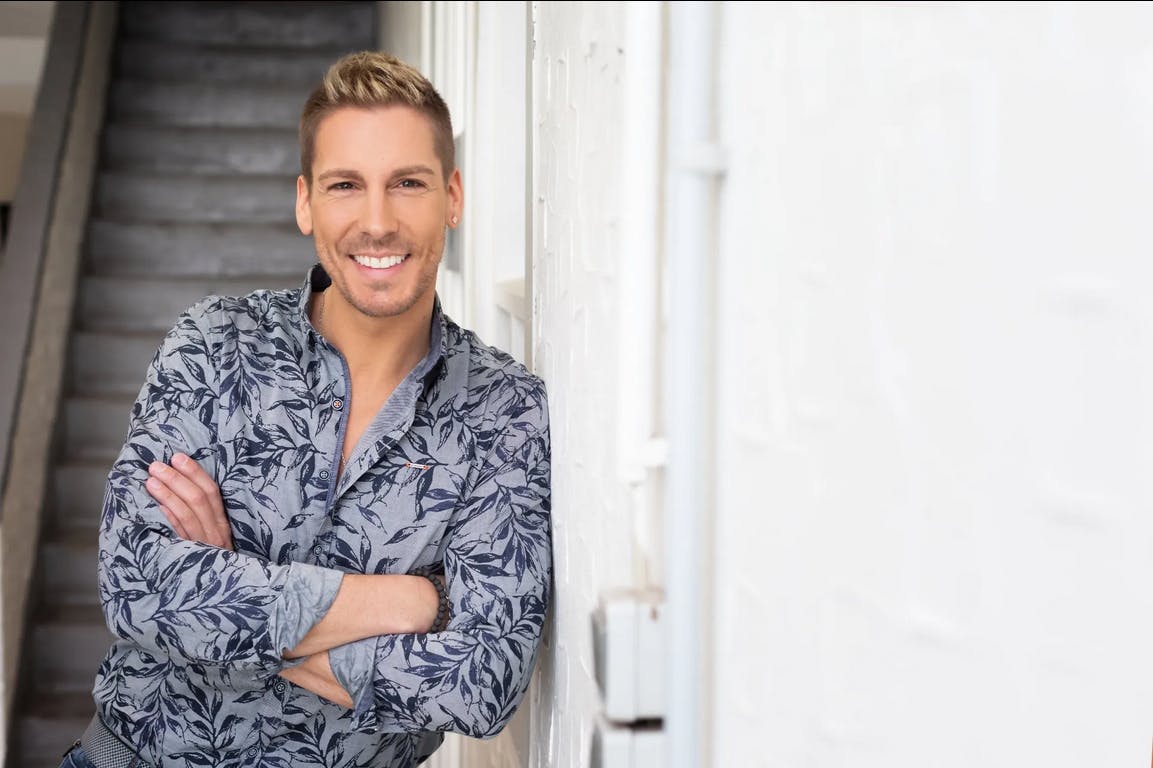 Junger Mann lehnt mit verschränkten Armen an einer weißen Wand vor einer Treppe. Er hat kurze, blond gesträhnte Haare, trägt ein hellgraues Hemd mit dunklem Blattmuster und lächelt freundlich in die Kamera.