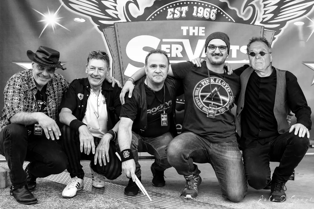 Fünf Männer knien nah nebeneinander vor einem großen Schild mit dem Schriftzug The Servats Est 1966. Alle schauen in die Kamera und lächeln. Der erste Mann von links trägt einen schwarzen Hut, ein gemustertes Hemd und dunkle Schuhe. Der zweite Mann trägt ein schwarzes Hemd über einem weißen T-Shirt und weiße Sneakers. Der dritte Mann trägt ein schwarzes T-Shirt mit Weste, eine schwarze Armbinde mit Schriftzug Bad to the Bone und hält Schlagzeugstöcke in der rechten Hand. Der vierte Mann trägt ein dunkles T-Shirt mit der Aufschrift Pink Floyd und eine graue Mütze sowie eine Brille. Der fünfte Mann trägt ein schwarzes T-Shirt, eine dunkle Weste und runde Sonnenbrillen. Alle wirken entspannt und freundlich. Unten rechts steht der Schriftzug John D. Grant Photography.