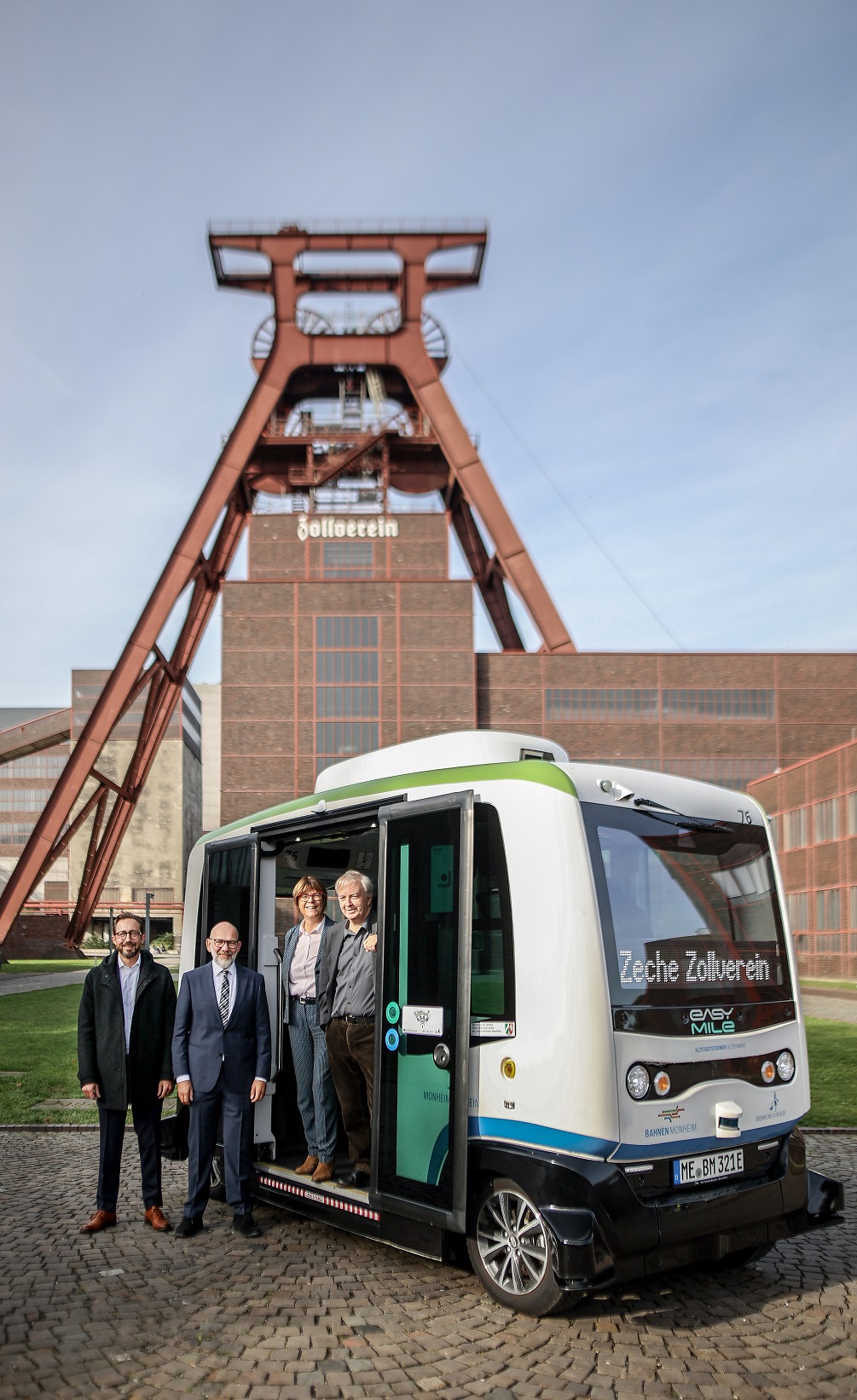 Vor dem bekannten Förderturm der Zeche Zollverein in Essen steht ein weiß-grüner autonomer Shuttlebus mit der Aufschrift Zeche Zollverein. Neben dem Bus auf dem Kopfsteinpflaster und teils im Einstieg stehend posieren vier Erwachsene für ein Foto, zwei davon befinden sich im Fahrzeug. Im Hintergrund ist das rote Stahlgerüst des Industriedenkmals sowie das markante Backsteingebäude mit dem Schriftzug Zollverein zu sehen. Die Szene verbindet moderne Mobilität mit dem historischen Ort der Industriekultur.