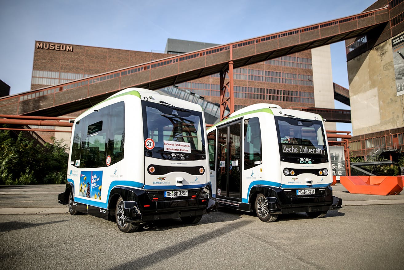 Das Bild zeigt zwei kleine autonome Shuttlebusse der Marke EasyMile auf dem Gelände der Zeche Zollverein in Essen. Die Busse sind weiß mit grünen und blauen Akzenten und stehen nebeneinander auf einer Straße vor der markanten Backsteinarchitektur des Ruhr Museums. Auf dem linken Bus ist ein Schild mit der Aufschrift Fahrzeug fährt autonom, Bitte Abstand halten zu sehen, während der rechte Bus als Ziel Zeche Zollverein anzeigt. Im Hintergrund sind rostrote Förderbänder und industrielle Gebäude des Weltkulturerbes sichtbar. Die Szene wirkt modern und zeigt die Verbindung von technischer Innovation mit historischer Industriekultur.