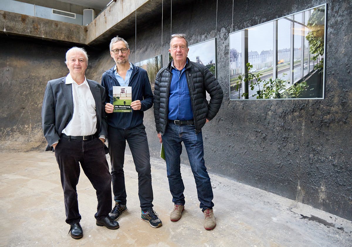Drei Männer stehen nebeneinander in einem Ausstellungsraum mit rauer, dunkler Wand und hängen Fotografien. Der Mann in der Mitte hält ein Buch mit dem Titel Die Wanderung in den Händen, auf dem ein Förderturm und eine Person auf einer Wiese zu sehen sind. Alle drei blicken freundlich in die Kamera. Links steht ein Mann mit weißem Haar im grauen Anzug, rechts ein Mann mit schwarzer Steppjacke und blauen Jeans. Die Atmosphäre ist sachlich und zugleich entspannt.