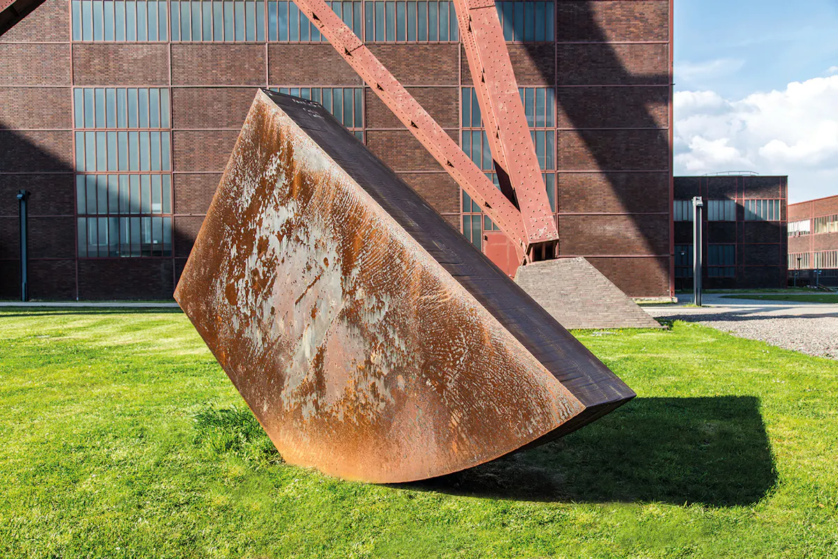 Große rostige Stahlskulptur mit schräg geneigter Fläche steht auf einer gepflegten Rasenfläche vor einem Backsteingebäude mit hohen Fenstern. Im Hintergrund ragen Teile eines rotbraunen Fördergerüsts in den Himmel, das zur Zeche Zollverein gehört. Die Szene ist bei sonnigem Wetter aufgenommen und zeigt deutliche Schatten auf dem Rasen.