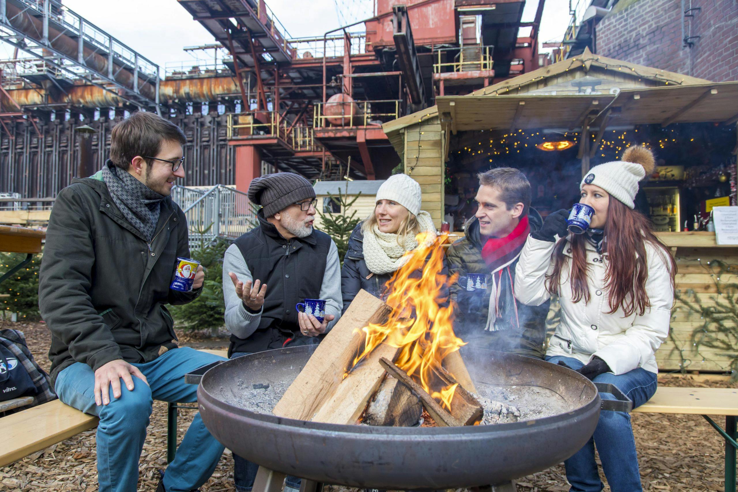 Fünf Personen in Winterkleidung sitzen um eine große Feuerschale mit loderndem Feuer und unterhalten sich fröhlich, während sie aus Tassen trinken. Die Szene spielt sich im Außenbereich eines weihnachtlich geschmückten Holzhäuschens ab, das Glühwein oder ähnliche Heißgetränke anbietet. Im Hintergrund ist ein altes Industriegebäude mit Stahlkonstruktion zu sehen, was auf einen Veranstaltungsort im industriellen Ambiente hindeutet. Es herrscht eine gemütliche, winterliche Stimmung.