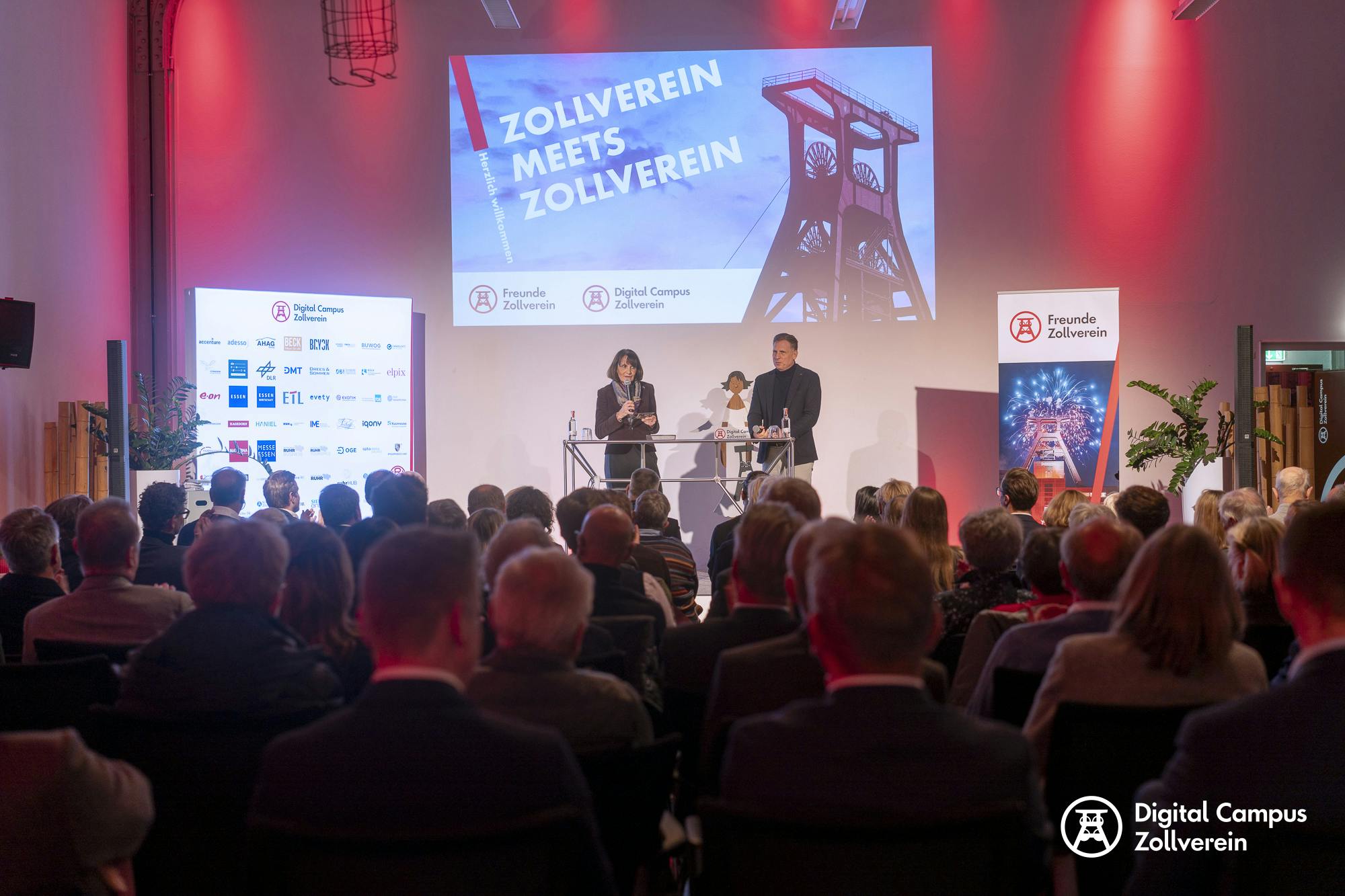 Auf einer Bühne stehen eine Frau und ein Mann hinter zwei Stehpulten und sprechen vor einem vollen Publikum. Über ihnen ist eine große Leinwand mit der Aufschrift Zollverein meets Zollverein sowie das Bild eines Förderturms zu sehen. Links und rechts der Bühne stehen Banner mit den Logos von Freunde Zollverein und Digital Campus Zollverein. Der Raum ist in warmes rotes Licht getaucht, die Besucherinnen und Besucher sitzen dicht beisammen und folgen aufmerksam dem Vortrag. Links auf der Bühne ist eine Sponsorentafel mit zahlreichen Logos zu sehen.
