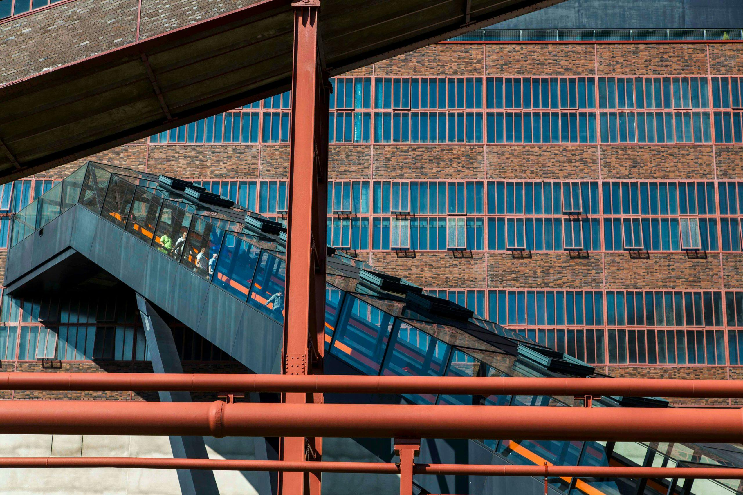 Außenaufnahme der beeindruckenden Rolltreppe zur ehemaligen Kohlenwäsche auf dem UNESCO-Welterbe Zollverein in Essen. Die verglaste Rolltreppe mit orangefarbenem Handlauf führt diagonal durch das Bild nach oben. Dahinter ist die Fassade eines großen Industriegebäudes mit gleichmäßigen Fensterreihen in Backsteinoptik zu sehen. Im Vordergrund verlaufen rostrote Stahlträger und Rohre, die an die industrielle Vergangenheit des Ortes erinnern. Einige Personen sind durch die Glasscheiben der Rolltreppe sichtbar.