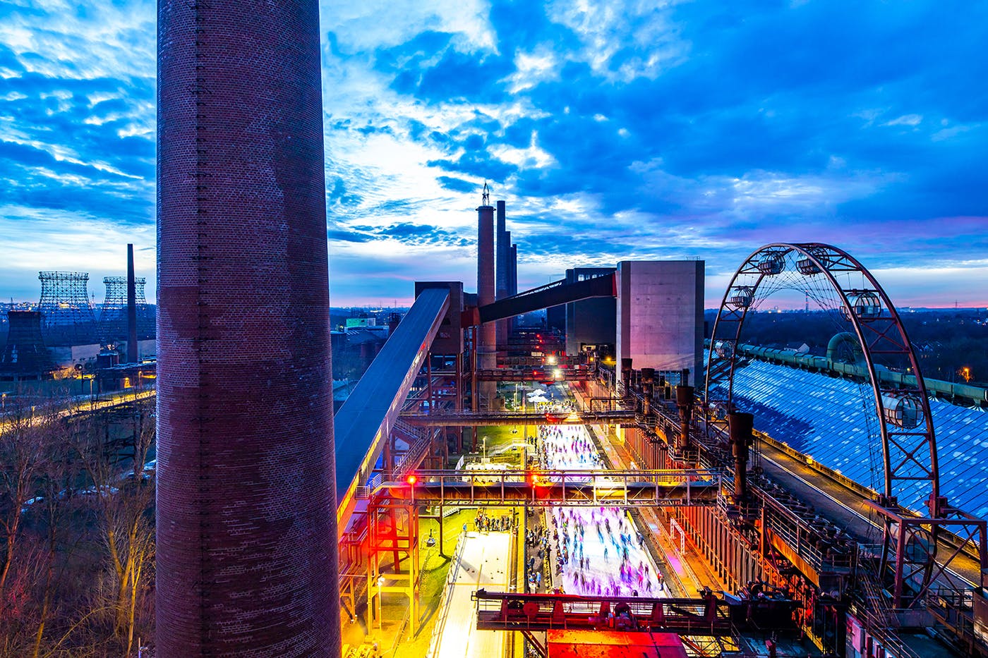 Abendliche Aufnahme eines ehemaligen Industriekomplexes mit einer hell beleuchteten Eislaufbahn im Zentrum, auf der viele Menschen unterwegs sind. Links im Bild erhebt sich ein hoher Schornstein aus rotem Backstein, rechts ist ein großes, halbrundes Gerüst eines stillgelegten Förderrads zu sehen. Im Hintergrund leuchten weitere Gebäude und ein dramatisch gefärbter Himmel in Blau- und Violetttönen kündigt die Dämmerung an.