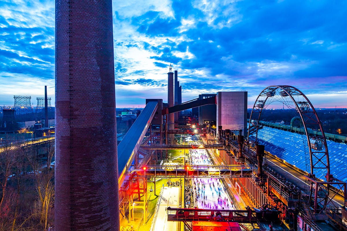 Abendliche Aufnahme eines ehemaligen Industriekomplexes mit einer hell beleuchteten Eislaufbahn im Zentrum, auf der viele Menschen unterwegs sind. Links im Bild erhebt sich ein hoher Schornstein aus rotem Backstein, rechts ist ein großes, halbrundes Gerüst eines stillgelegten Förderrads zu sehen. Im Hintergrund leuchten weitere Gebäude und ein dramatisch gefärbter Himmel in Blau- und Violetttönen kündigt die Dämmerung an.